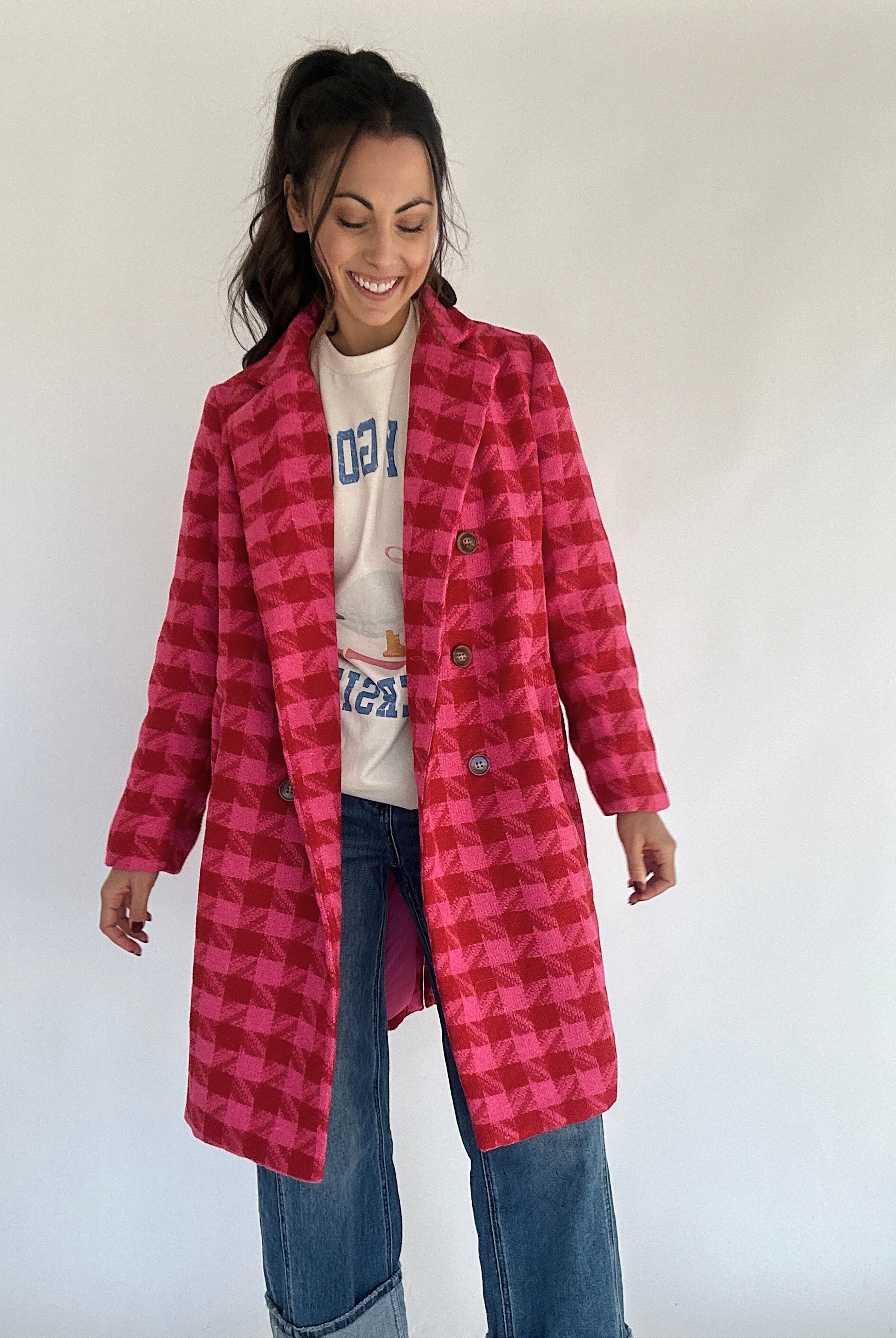 A person standing and smiling, wearing a hot pink and red houndstooth tweed coat with a double button front and lapel, paired with blue jeans.