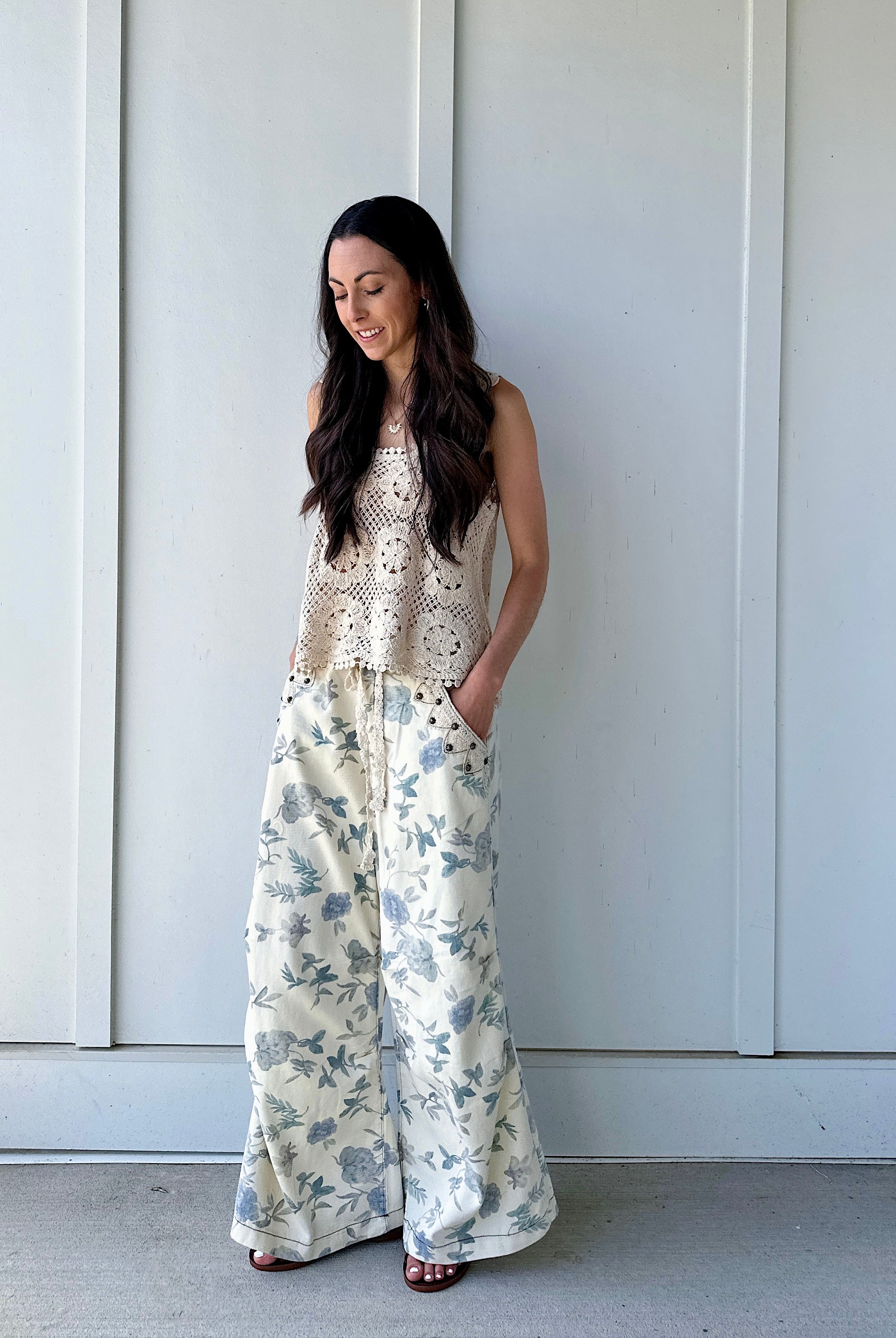 Woman wearing a crochet top and floral wide-leg pants with hands in pockets against a white wall.