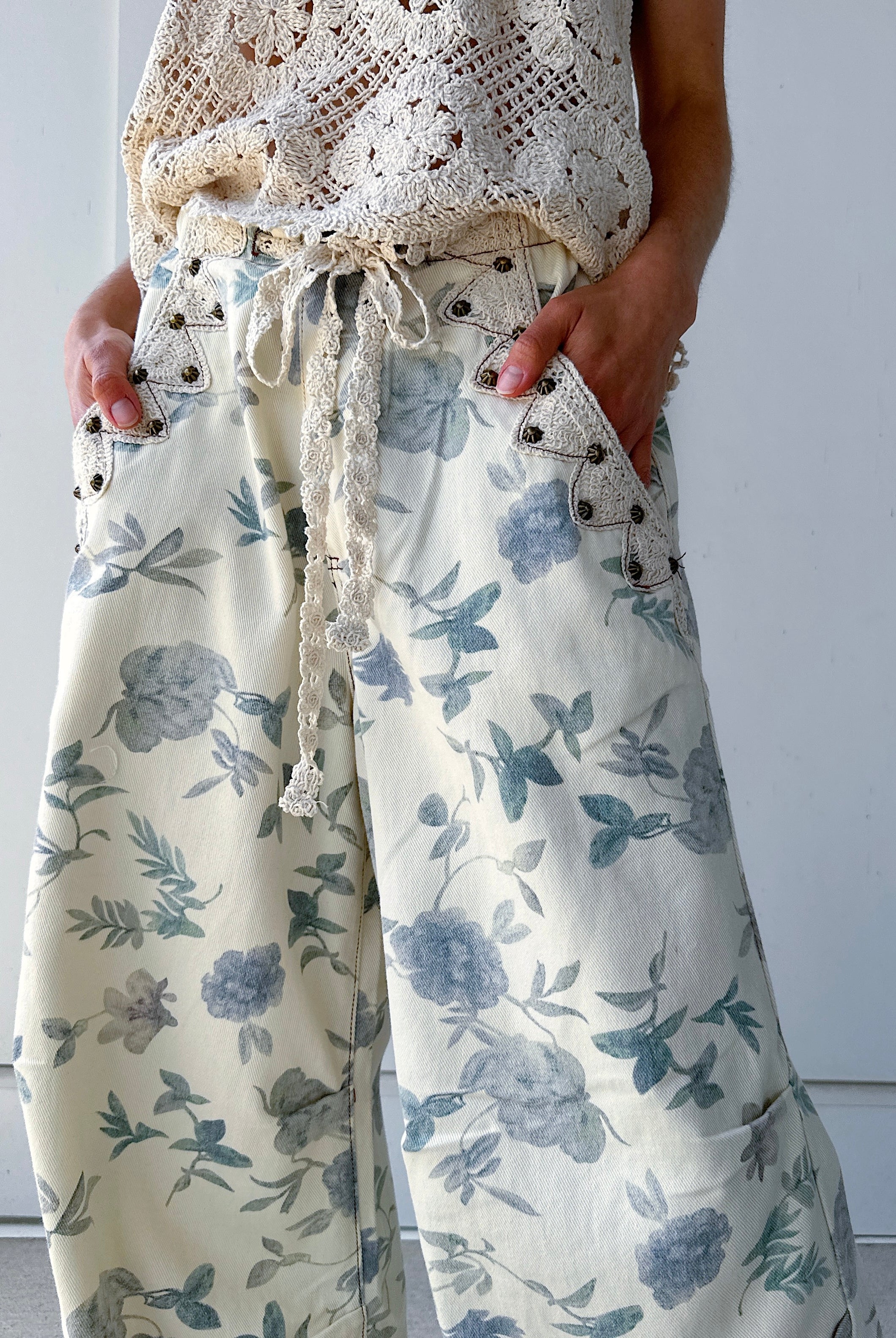Person wearing a white lace top and floral pants against a plain background with hands in pockets that are studded and lined by crochet trim