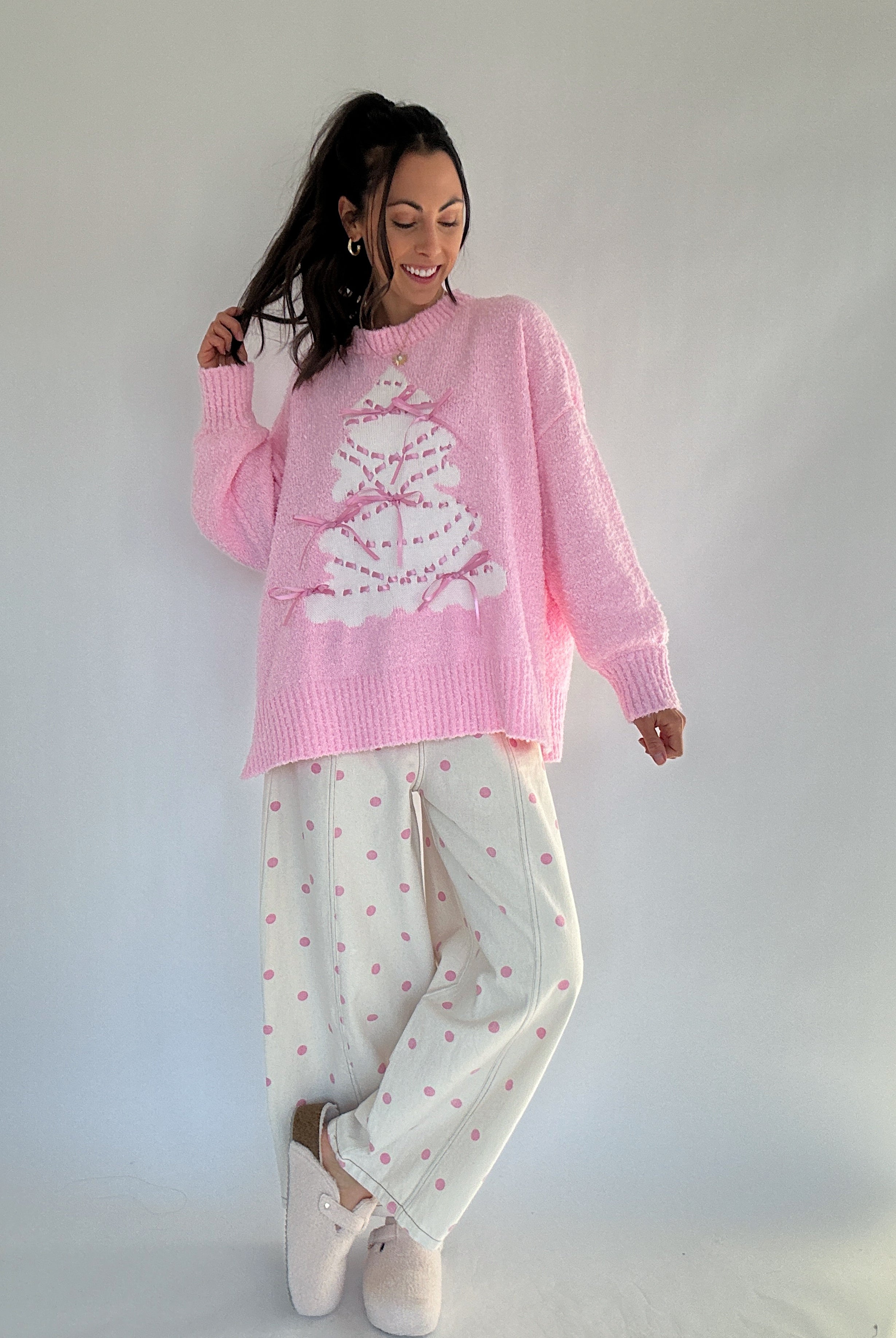 Woman wearing a pink sweater with a white tree design and pink polka dot pants against a white background