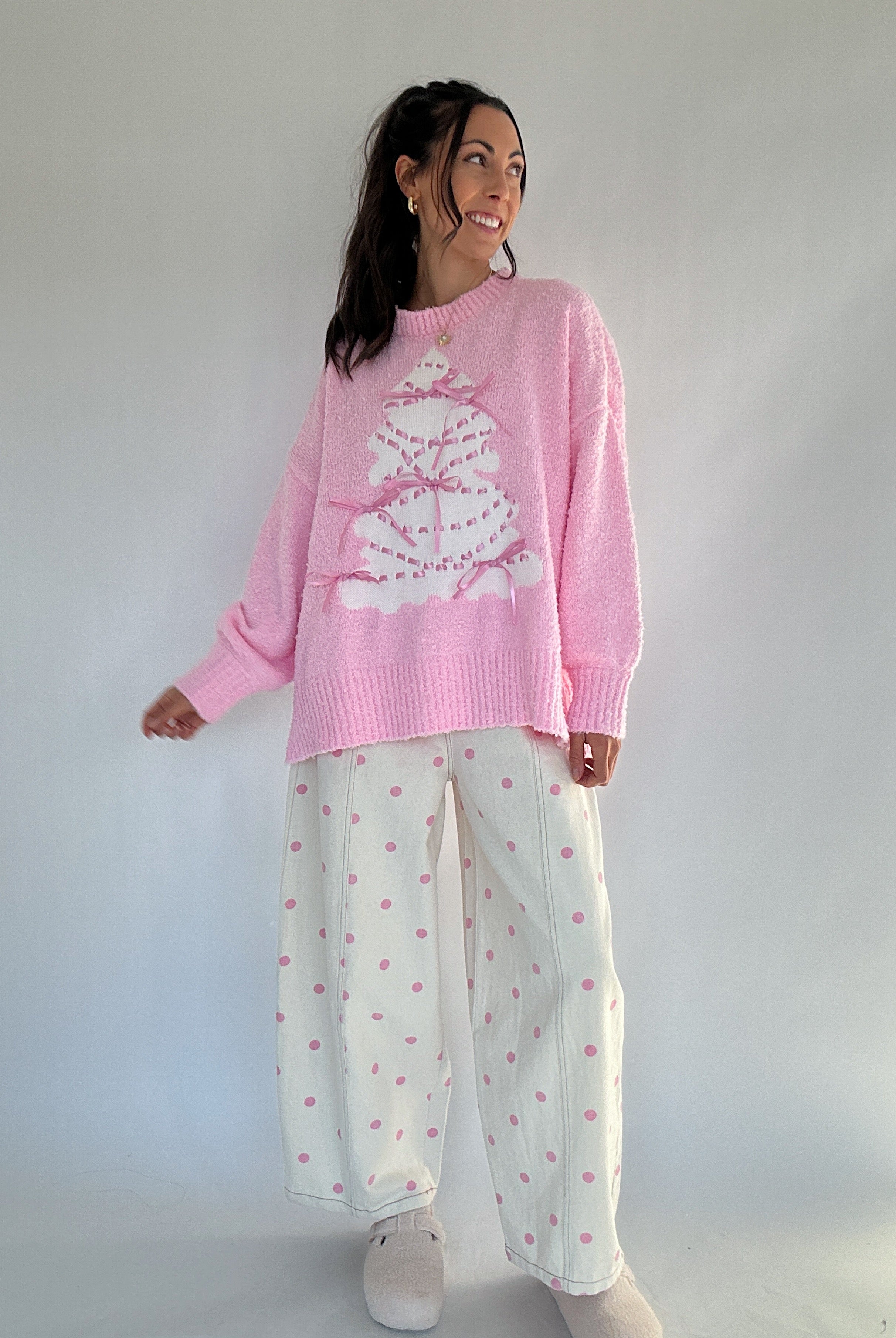 Woman wearing a pink sweater with a white tree design and white polka dot pants against a plain background