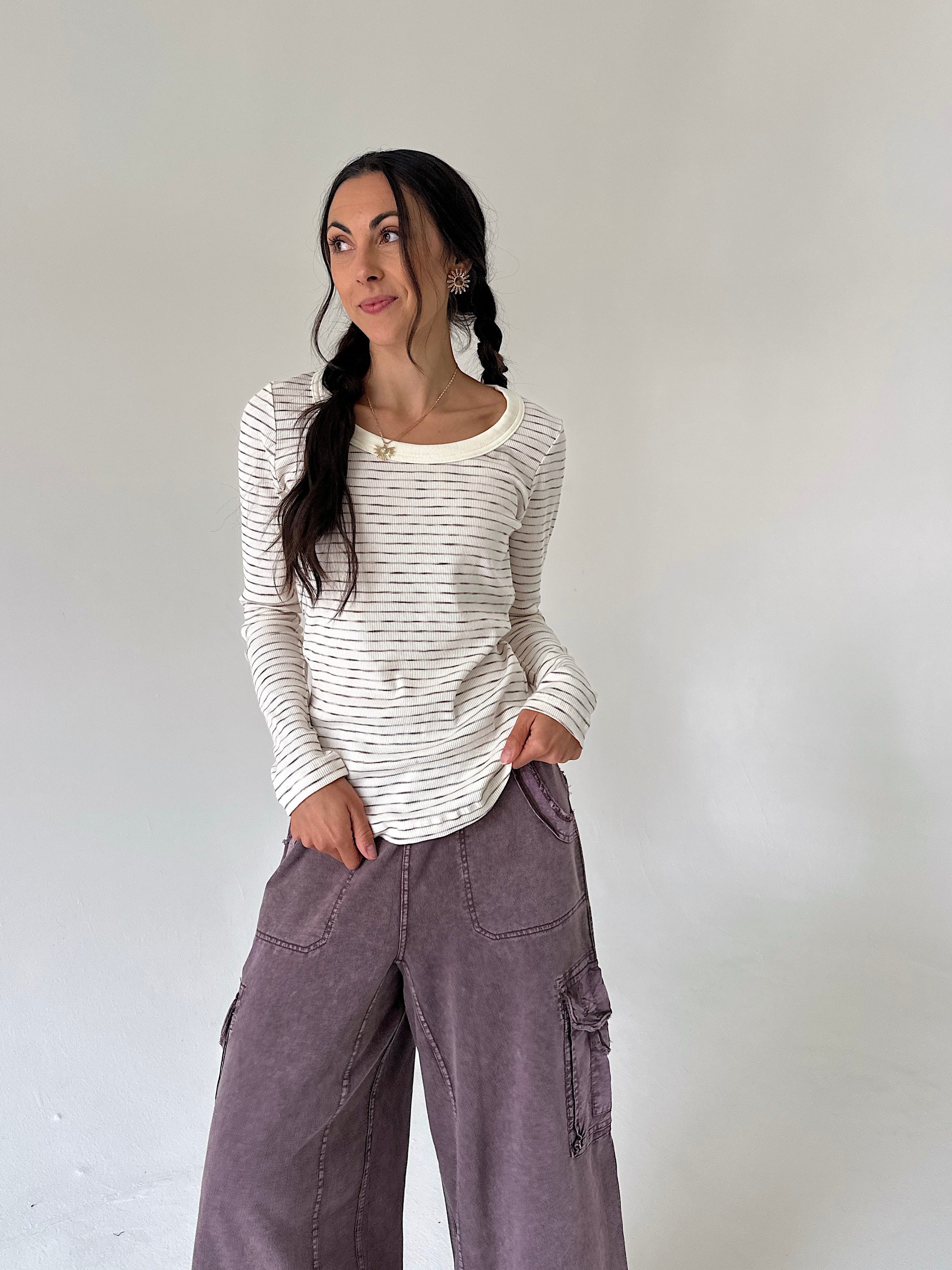 A woman with dark hair in two braids stands against a plain light gray wall, wearing a cream long-sleeve top with thin dark horizontal stripes and wide-leg mauve cargo pants.