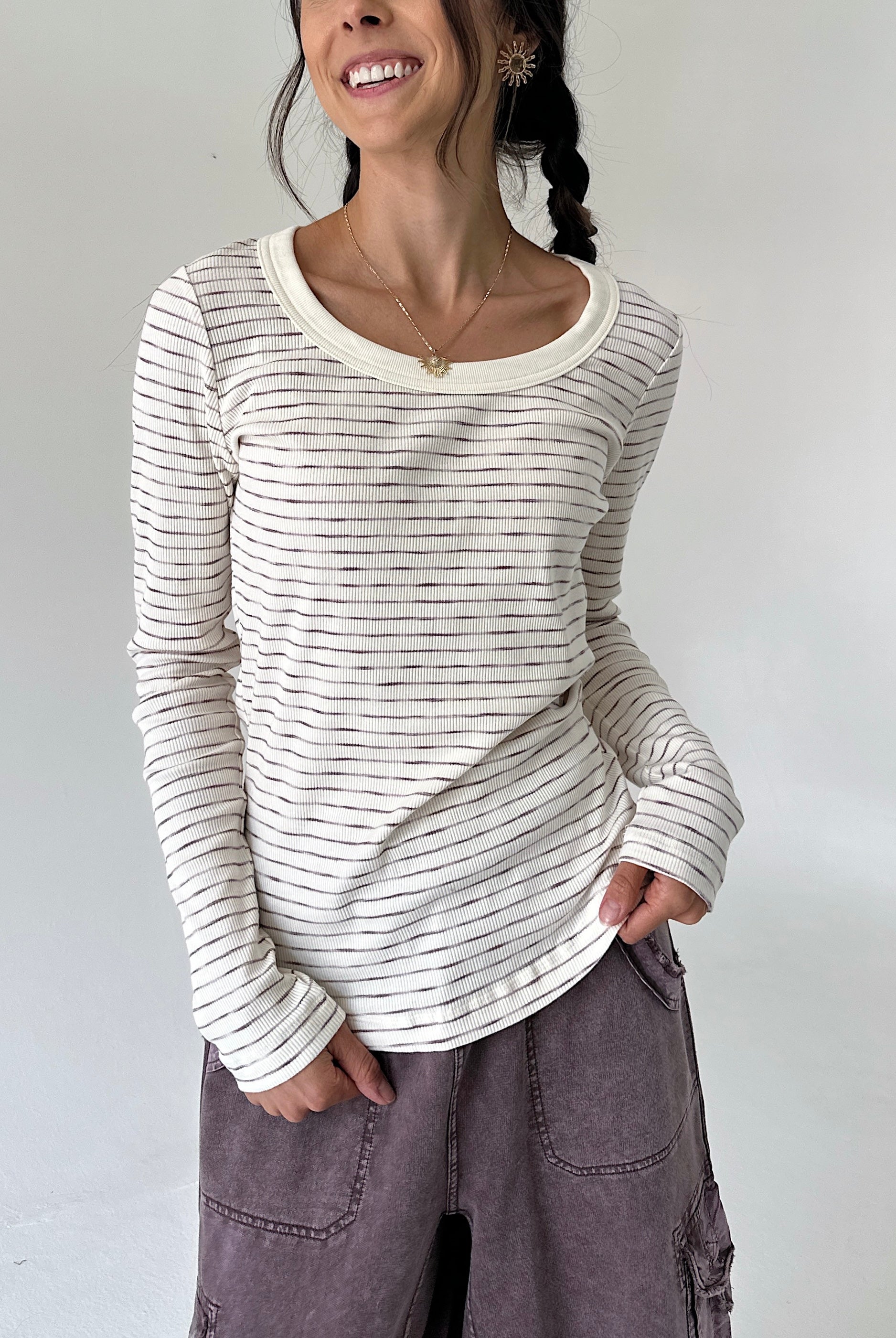 A woman wearing a cream-colored long-sleeve ribbed top with thin dark horizontal stripes and a scoop neckline, paired with dusty purple cargo pants, against a plain light gray background.