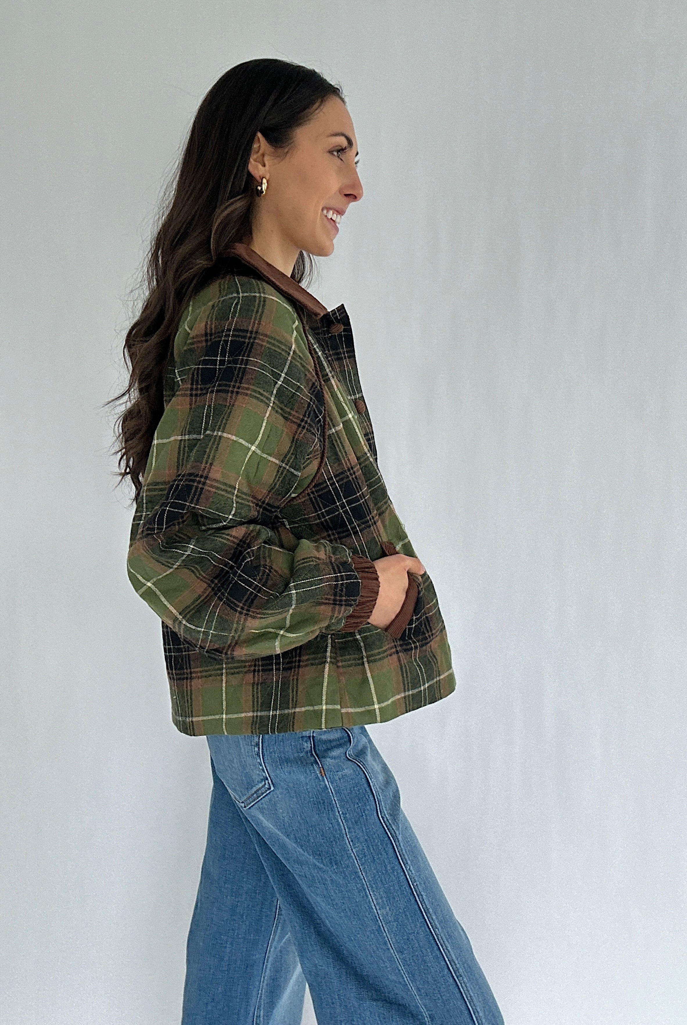 Woman wearing a green plaid jacket and blue jeans against a white background from side with hands in pockets