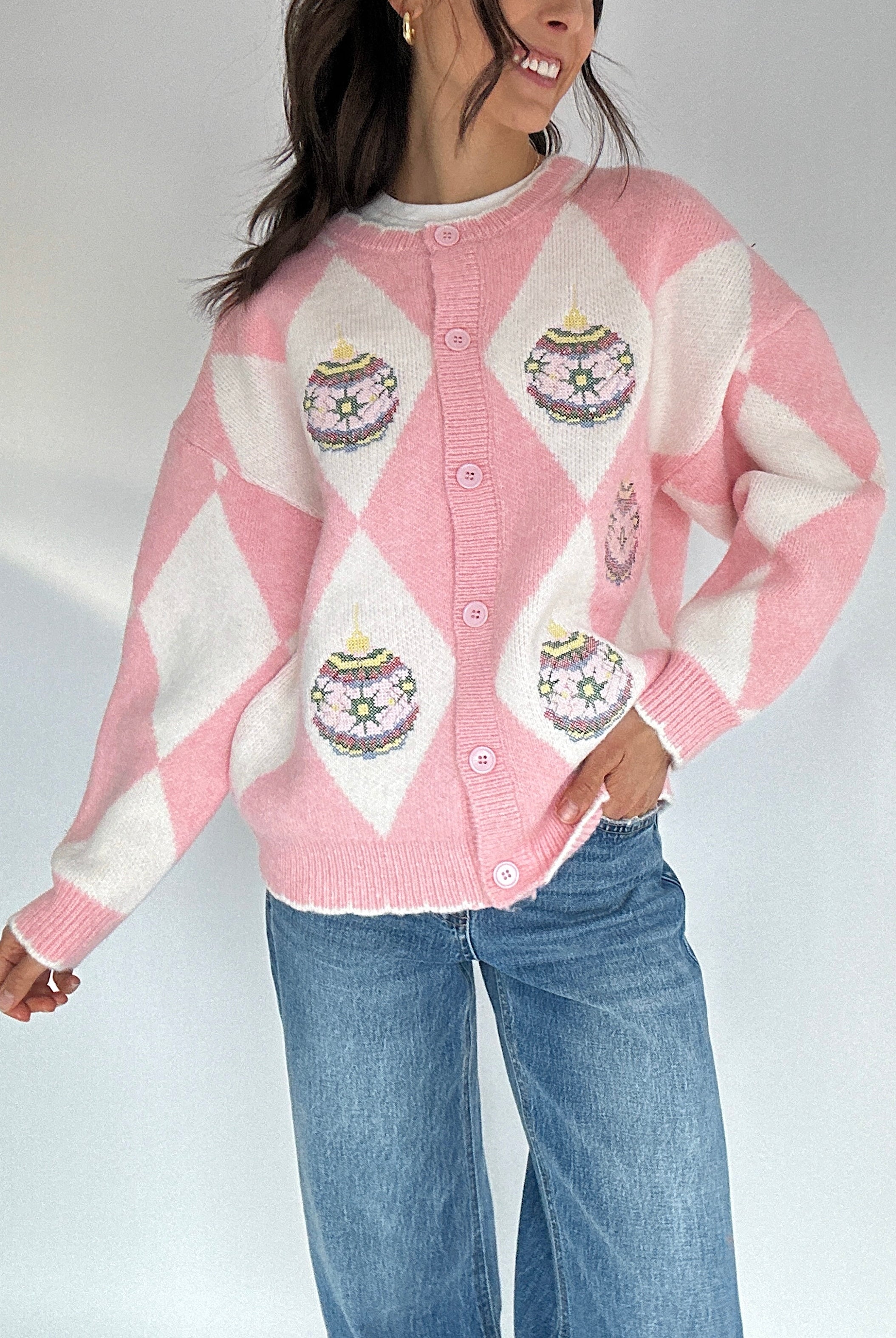 Person wearing a pink and white argyle and ornament patterned cardigan buttoned with blue jeans on a plain background