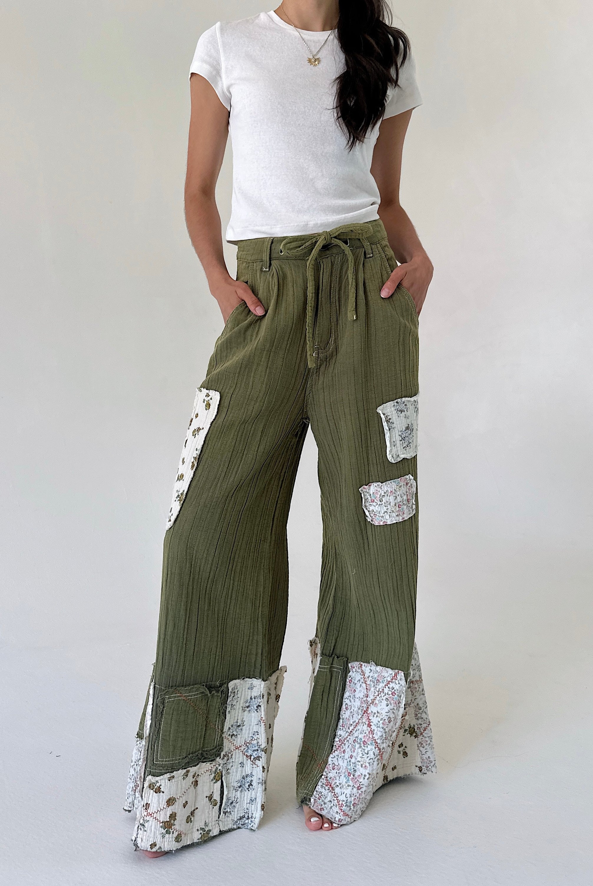 A model wearing olive green wide-leg patchwork pants with floral patterned inserts and a drawstring waist, paired with a white t-shirt against a plain light background.