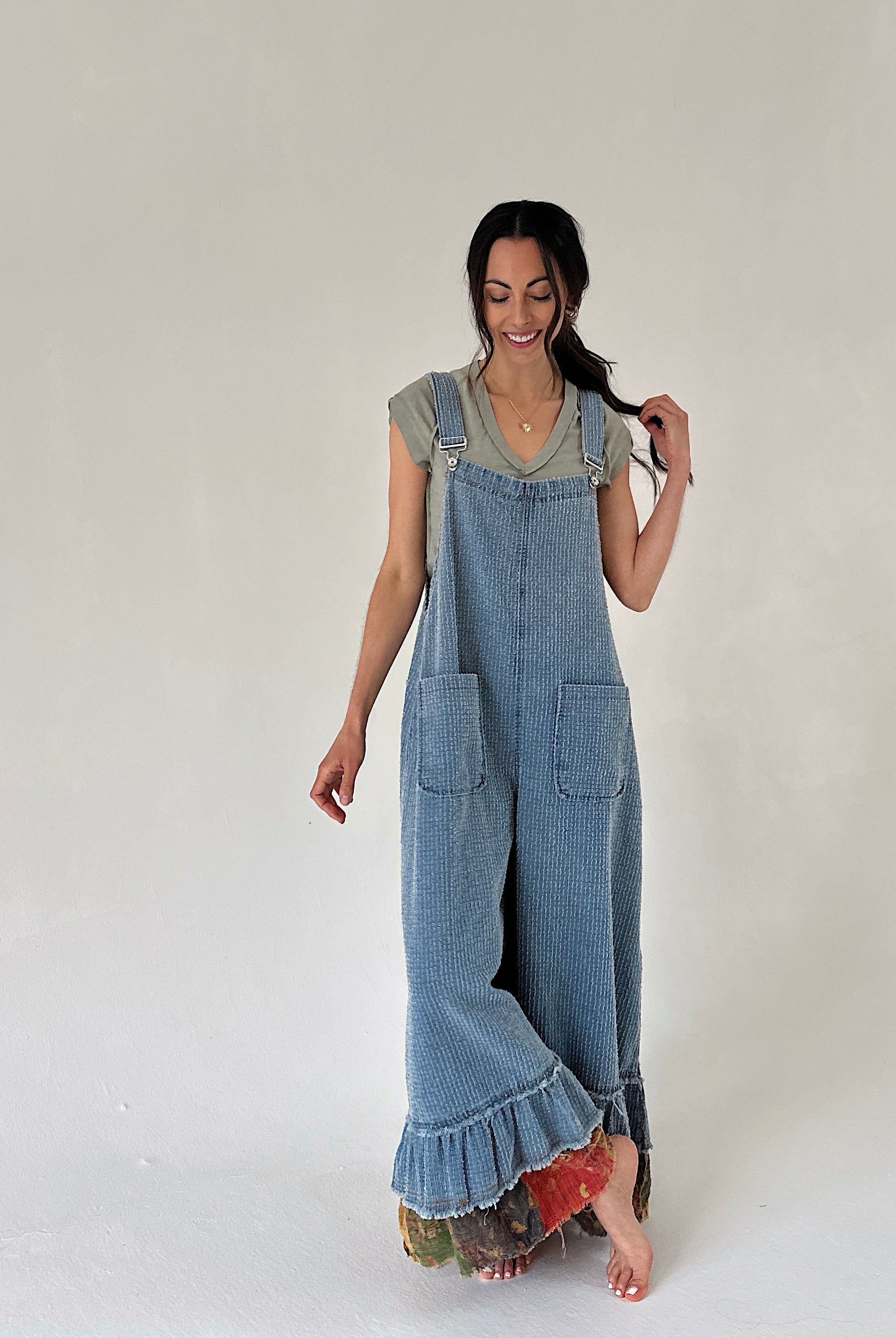 Woman wearing a denim overalls with a ruffle hem and legs crossed against a plain background