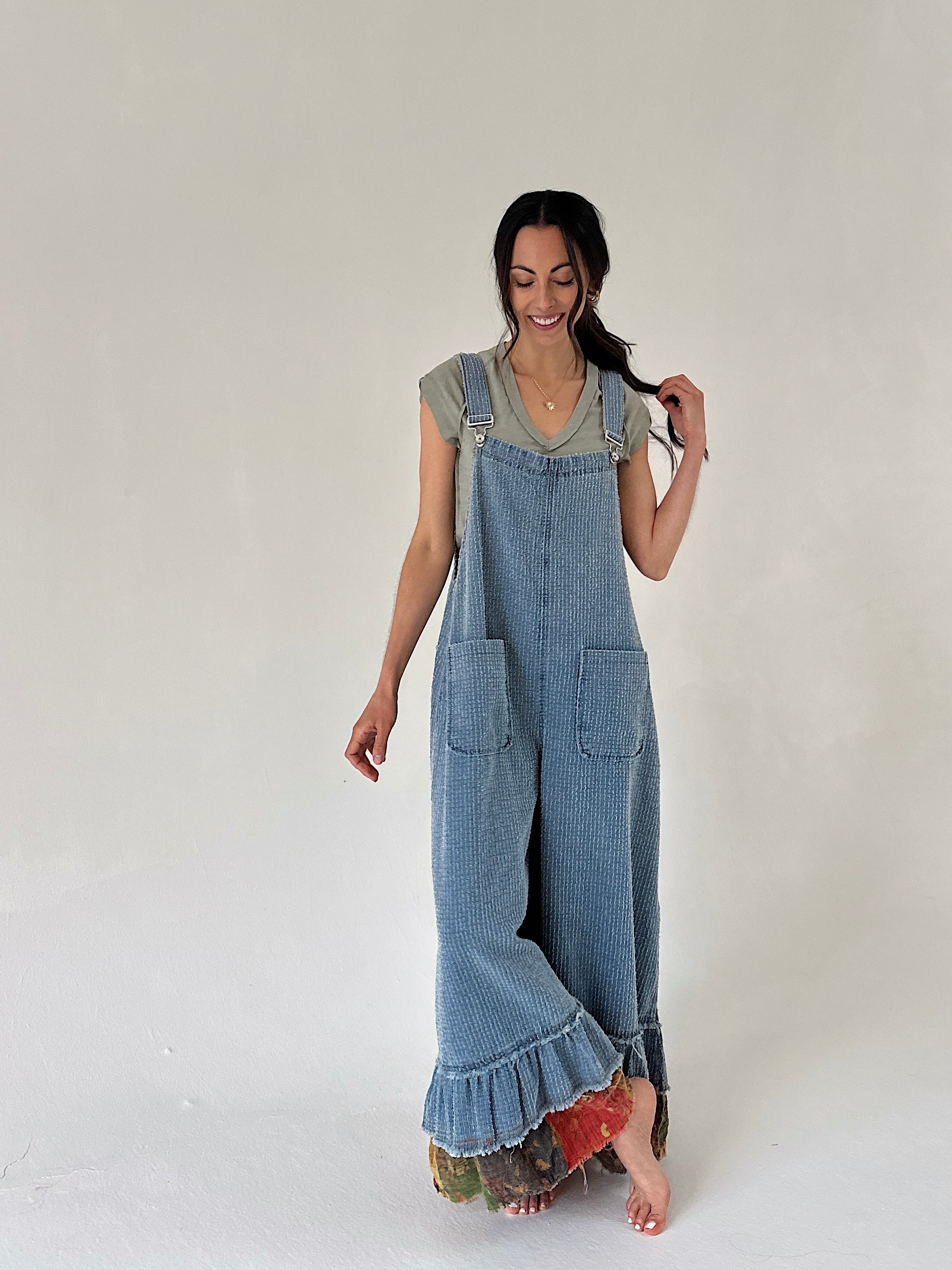 Woman wearing a denim overalls with a ruffle hem and legs crossed against a plain background