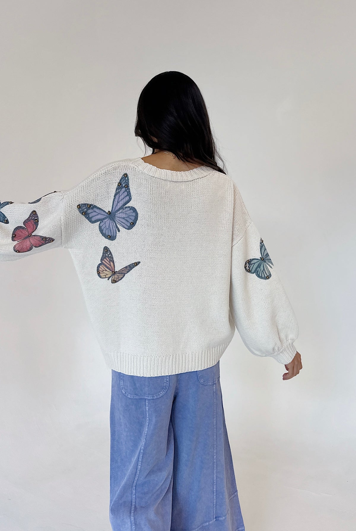 A person shown from behind wearing a cream knit sweater with blue, pink, and brown butterfly prints on the sleeves and back, paired with light blue wide-leg pants, against a plain off-white background.