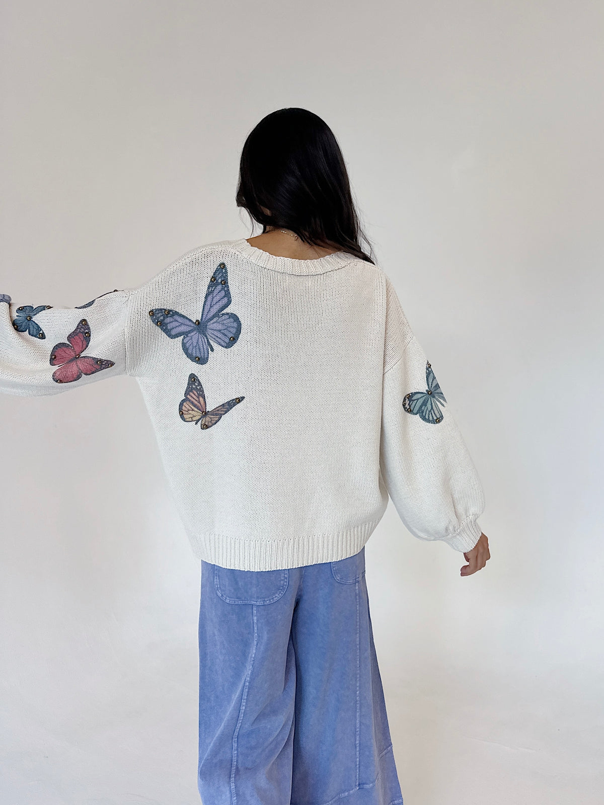 A person shown from behind wearing a cream knit sweater with blue, pink, and brown butterfly prints on the sleeves and back, paired with light blue wide-leg pants, against a plain off-white background.
