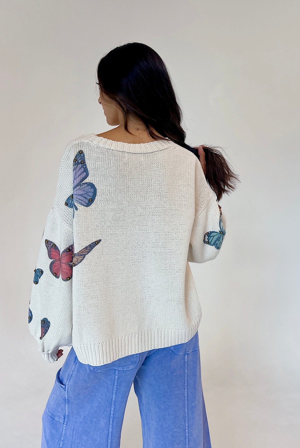 A woman viewed from behind wearing a cream-colored knit sweater with colorful butterfly prints on the back and sleeves, paired with light blue jeans, against a plain off-white wall.