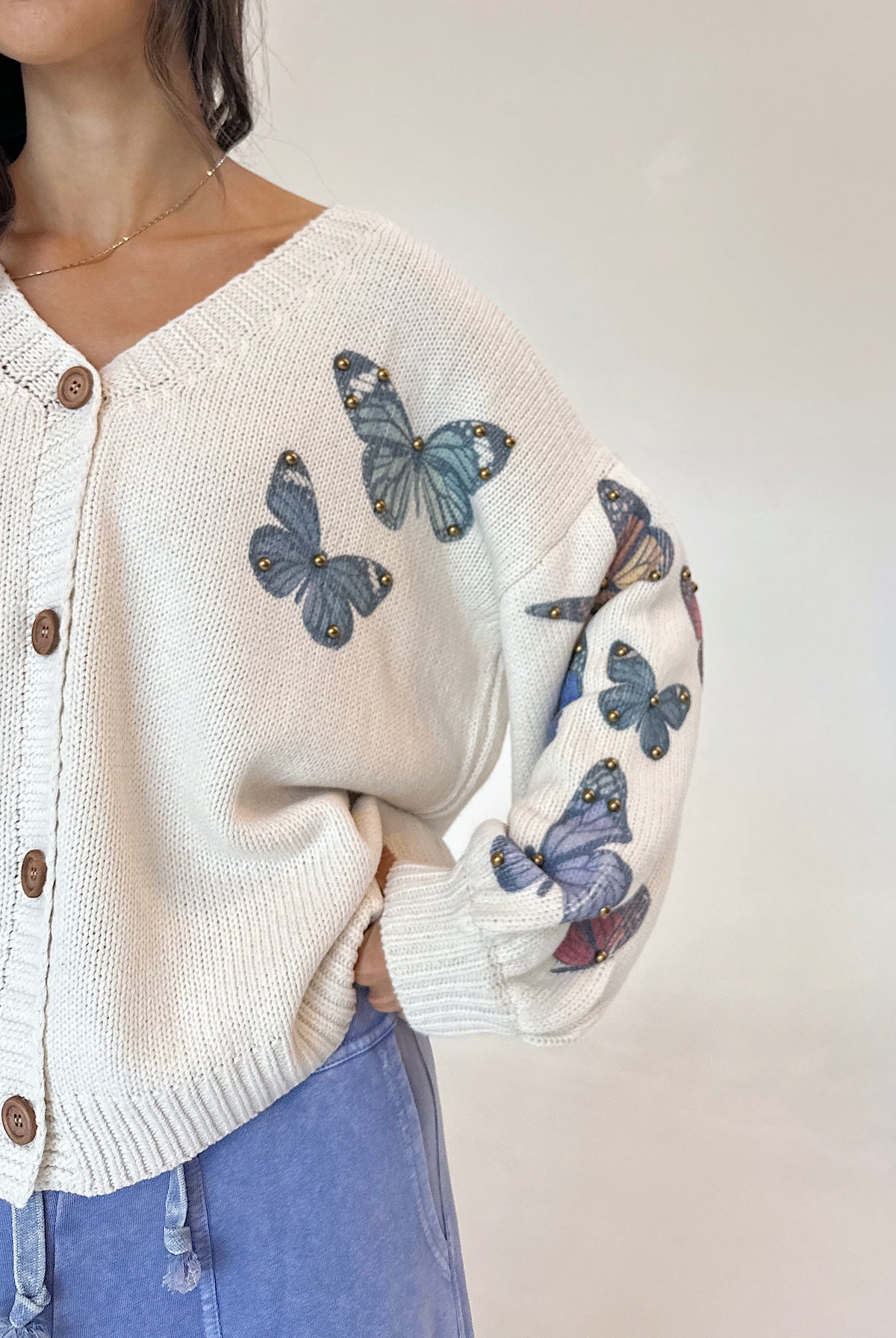 A close-up of a person wearing a cream knit cardigan with embroidered blue butterflies on the front and sleeves, paired with light blue jeans against a plain light background.