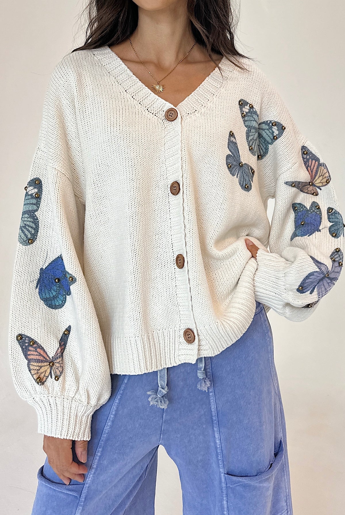 A woman wearing a cream-colored knit cardigan with blue and orange butterfly prints on the sleeves, paired with light blue pants, against a neutral beige background.