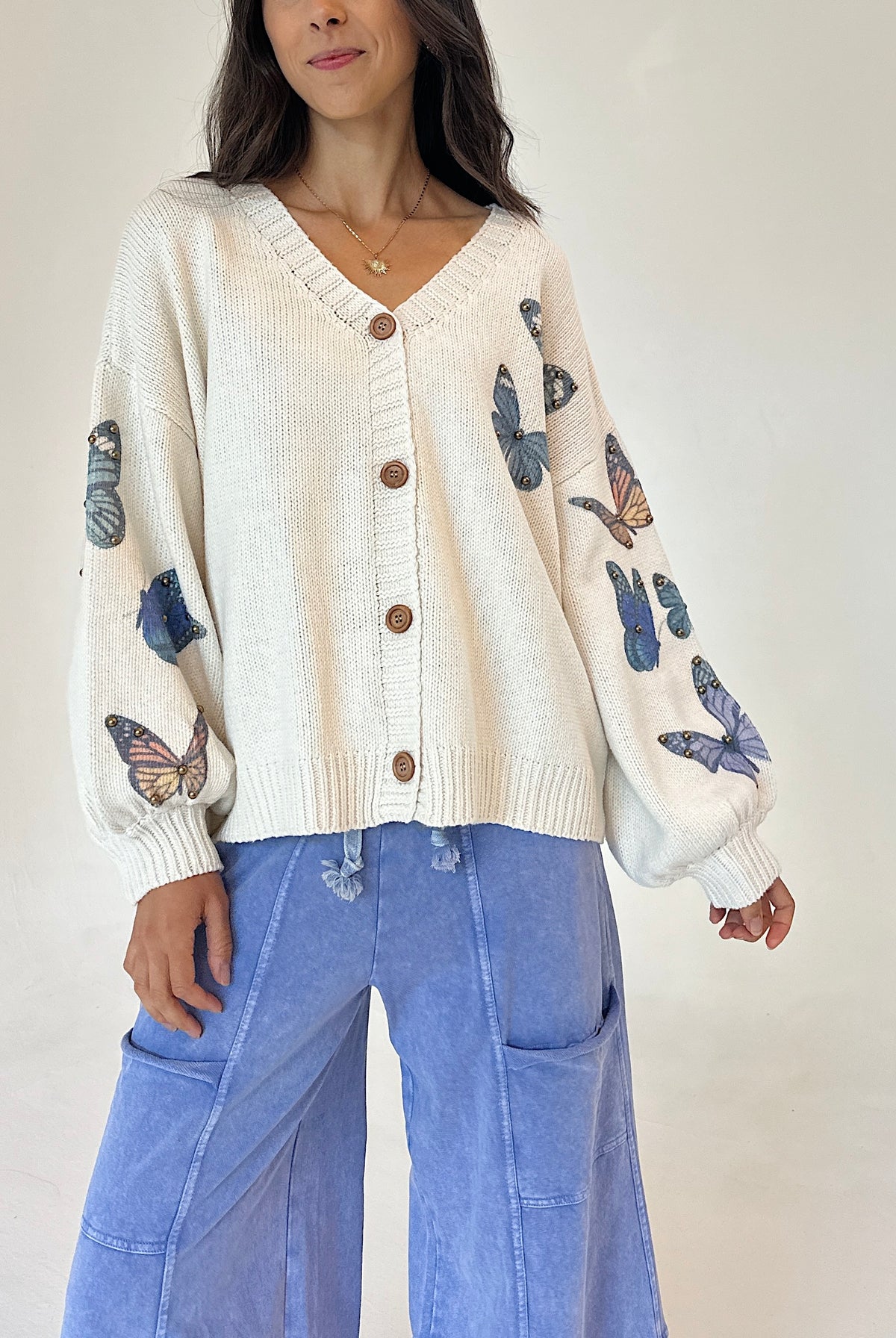 A woman wearing a cream-colored knit cardigan with brown buttons and blue butterfly prints on the sleeves, paired with light blue wide-leg pants, against a plain off-white background.