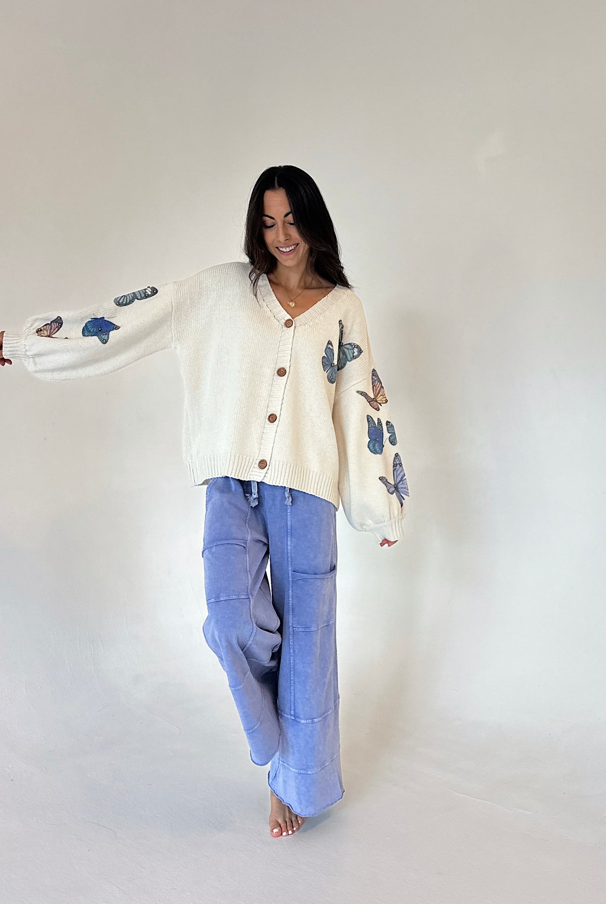 A woman modeling a cream-colored knit cardigan with blue butterfly embroidery on the sleeves, paired with light blue wide-leg pants, against a plain off-white studio background.