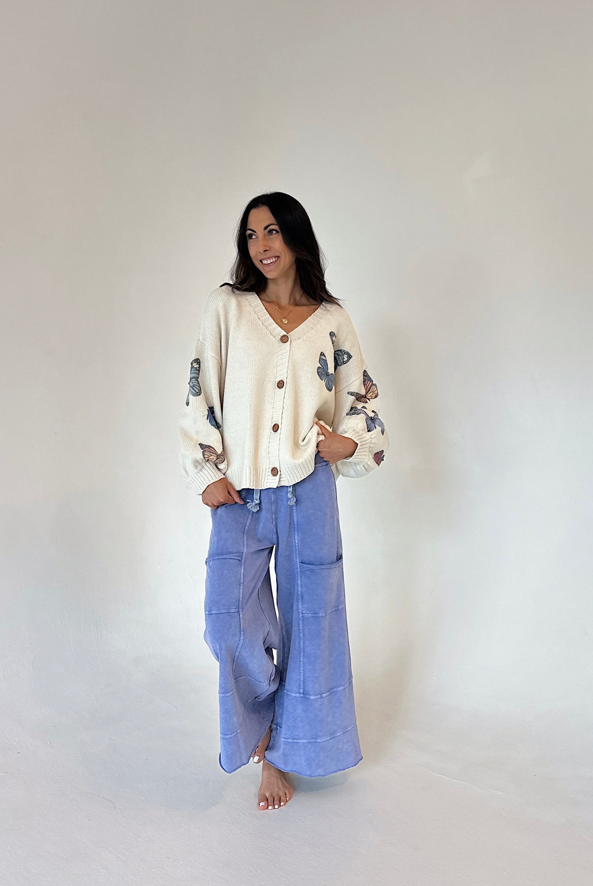 A woman stands barefoot against a plain light gray studio background, wearing a cream cardigan with blue butterfly embroidery on the sleeves and blue wide-leg pants.