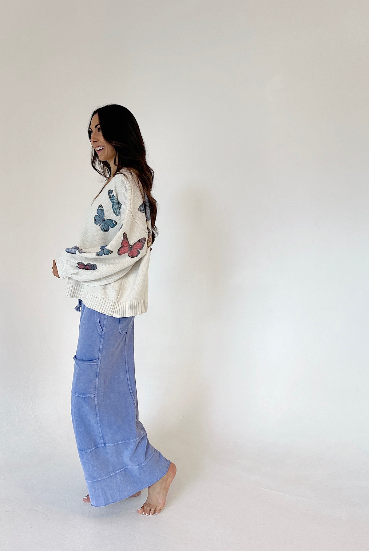 A woman stands barefoot against a plain white studio background, wearing a cream knit sweater with colorful butterfly prints and blue wide leg pants from side.
