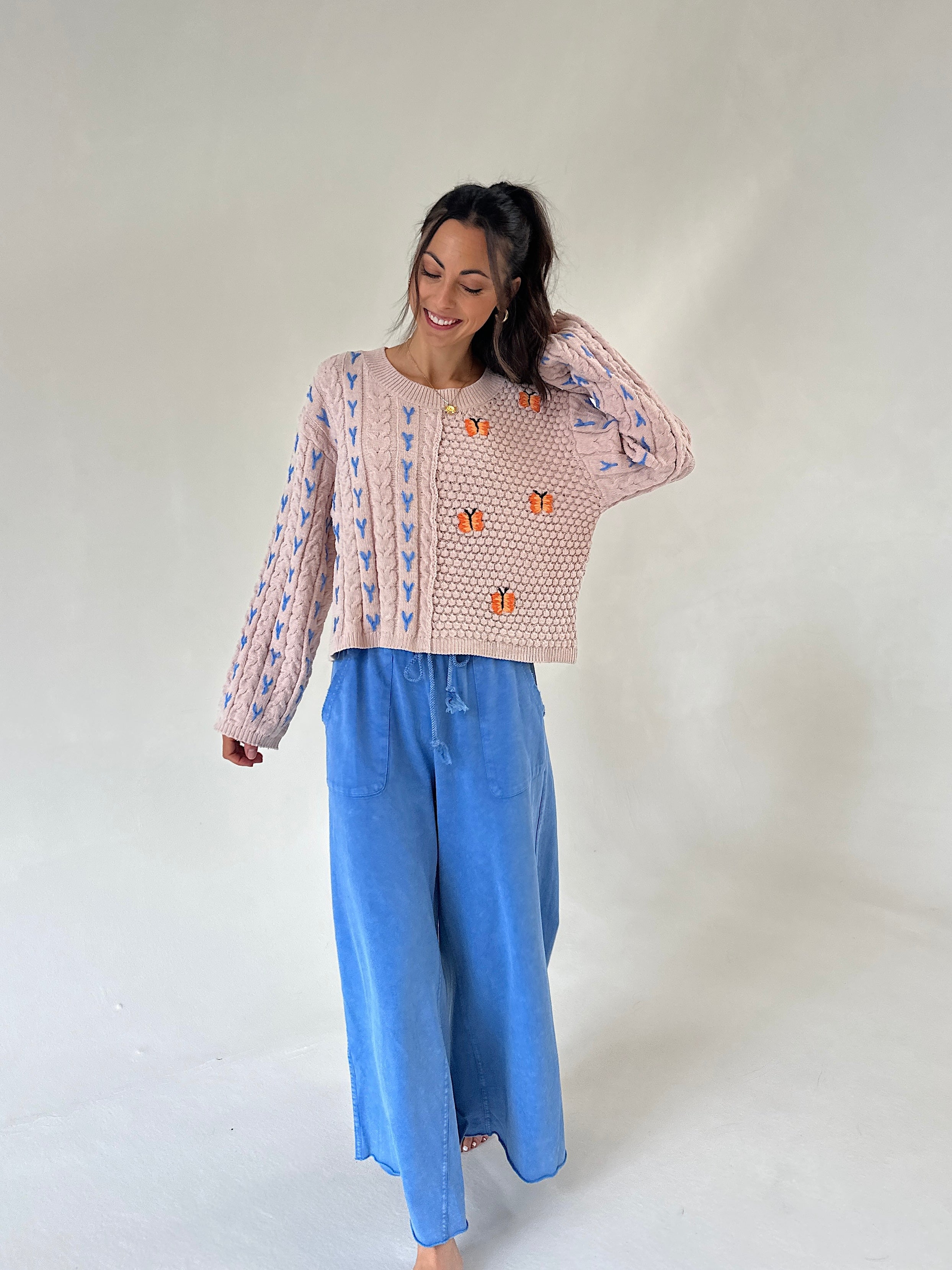 A woman modeling a taupe knit sweater with blue and orange embroidered patterns, paired with wide-leg blue pants, against a plain light gray studio background.