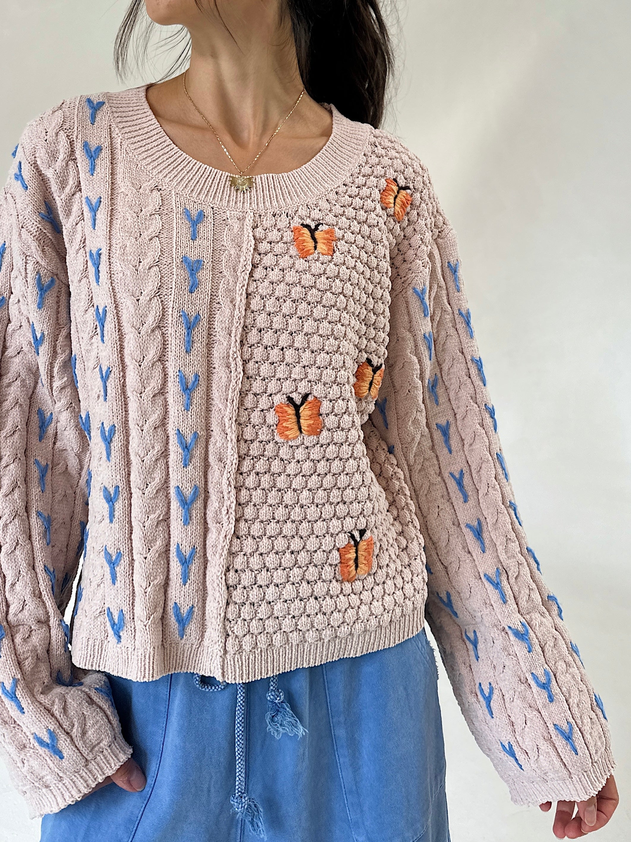 A woman wearing a taupe cable-knit sweater with blue patterns and orange butterfly appliqués, paired with blue pants against a plain white background.