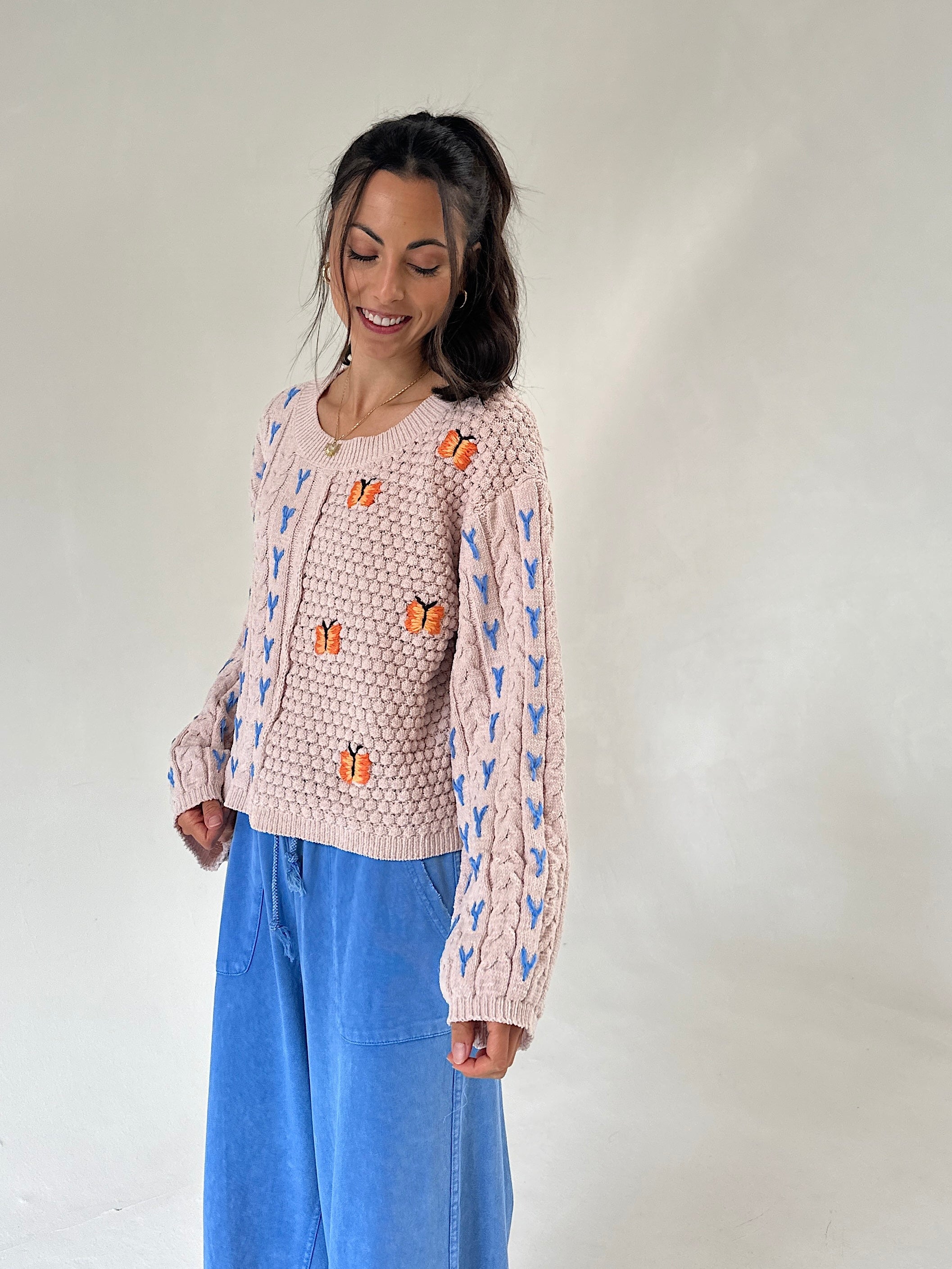 A woman wearing a taupe textured knit sweater with blue patterns and orange butterfly appliqués, paired with wide-leg blue pants, standing against a plain light gray studio background.