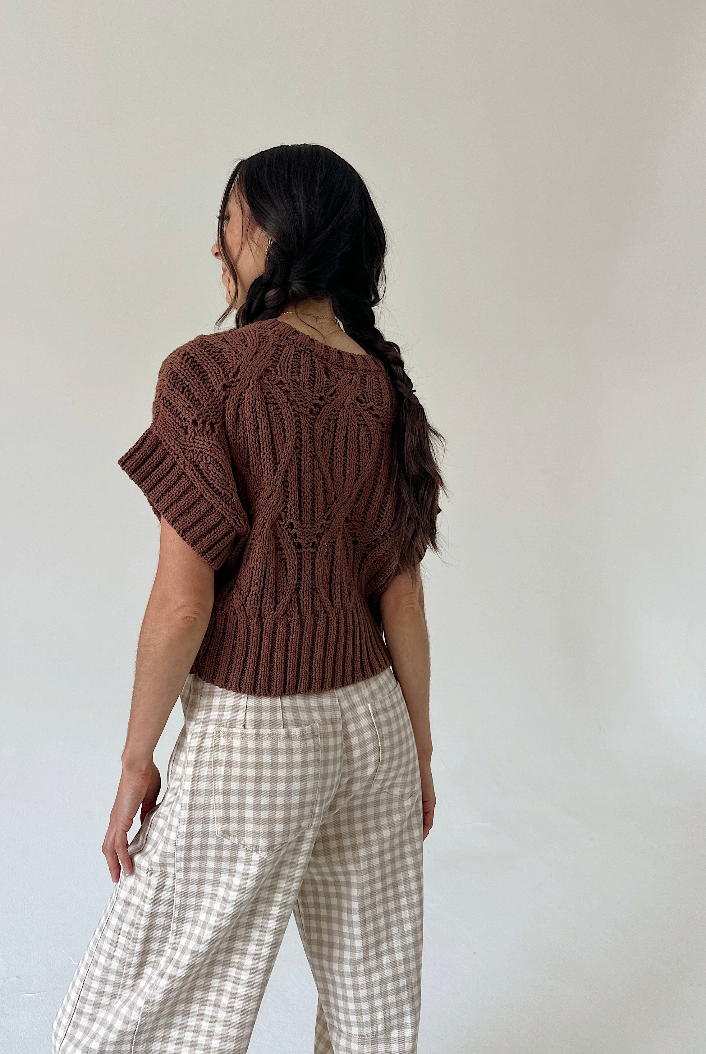 A woman shown from the back wearing a brown short-sleeve knit top and beige-and-white gingham pants against a plain off-white wall.