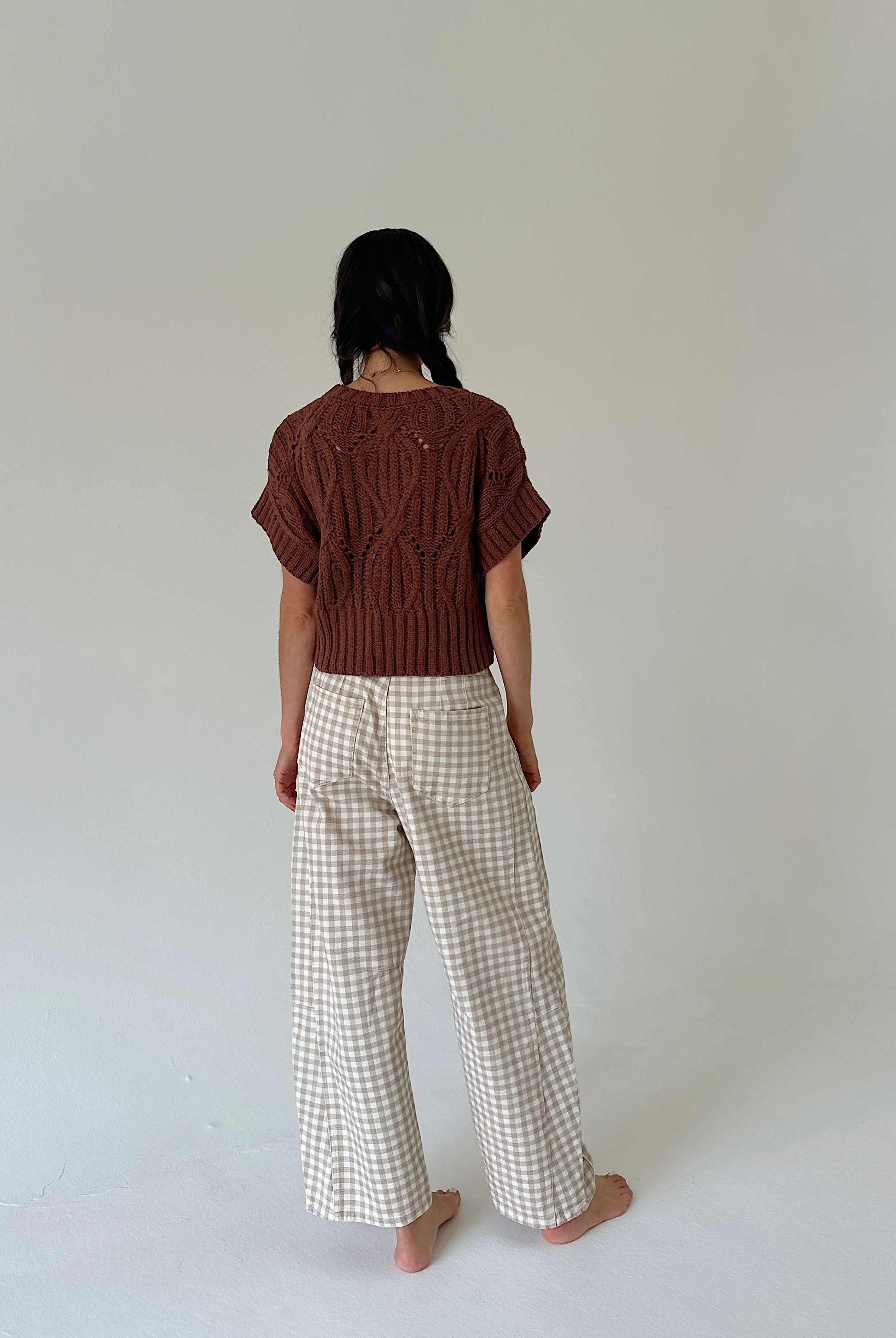 A person standing with their back to the camera, wearing a brown short-sleeve knit top and beige-and-white gingham wide-leg trousers, against a plain light gray studio background.