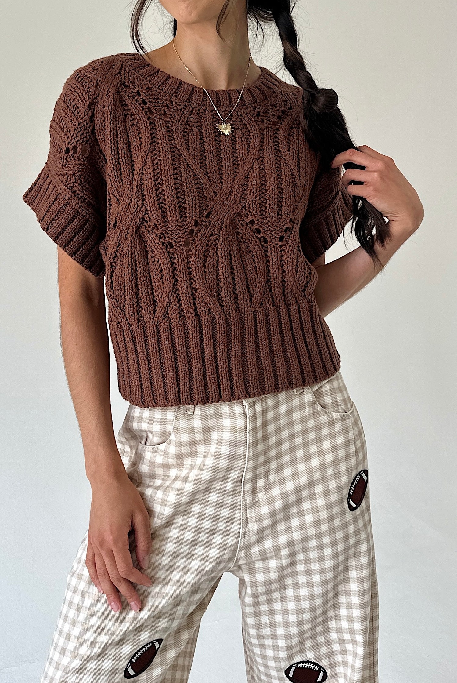 A person wearing a brown short-sleeve cable-knit sweater and beige-and-white gingham pants with football patch details, photographed against a plain light background.