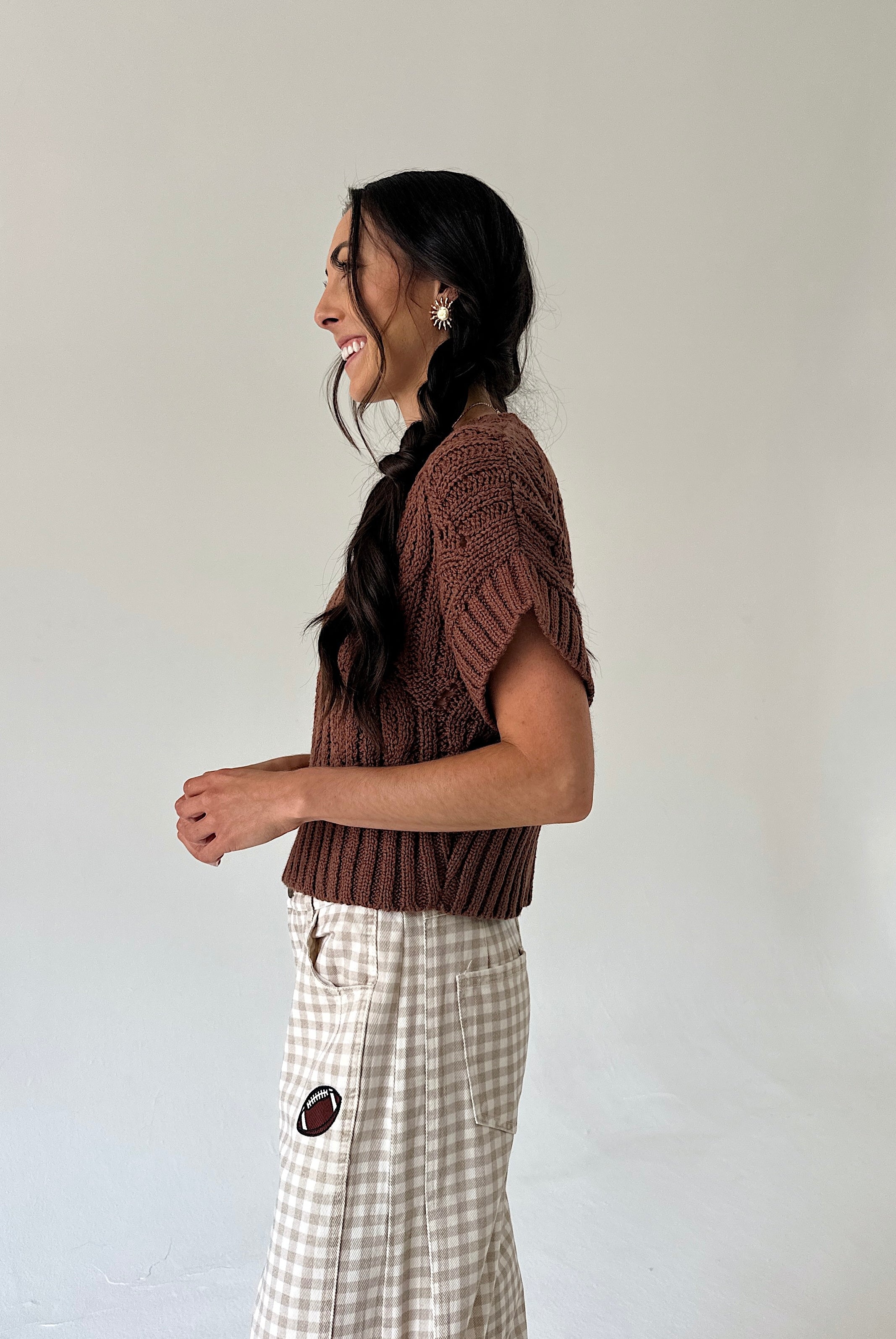 A woman in profile wearing a brown short-sleeve knit top and a beige-and-white gingham pant, standing against a plain light gray wall.