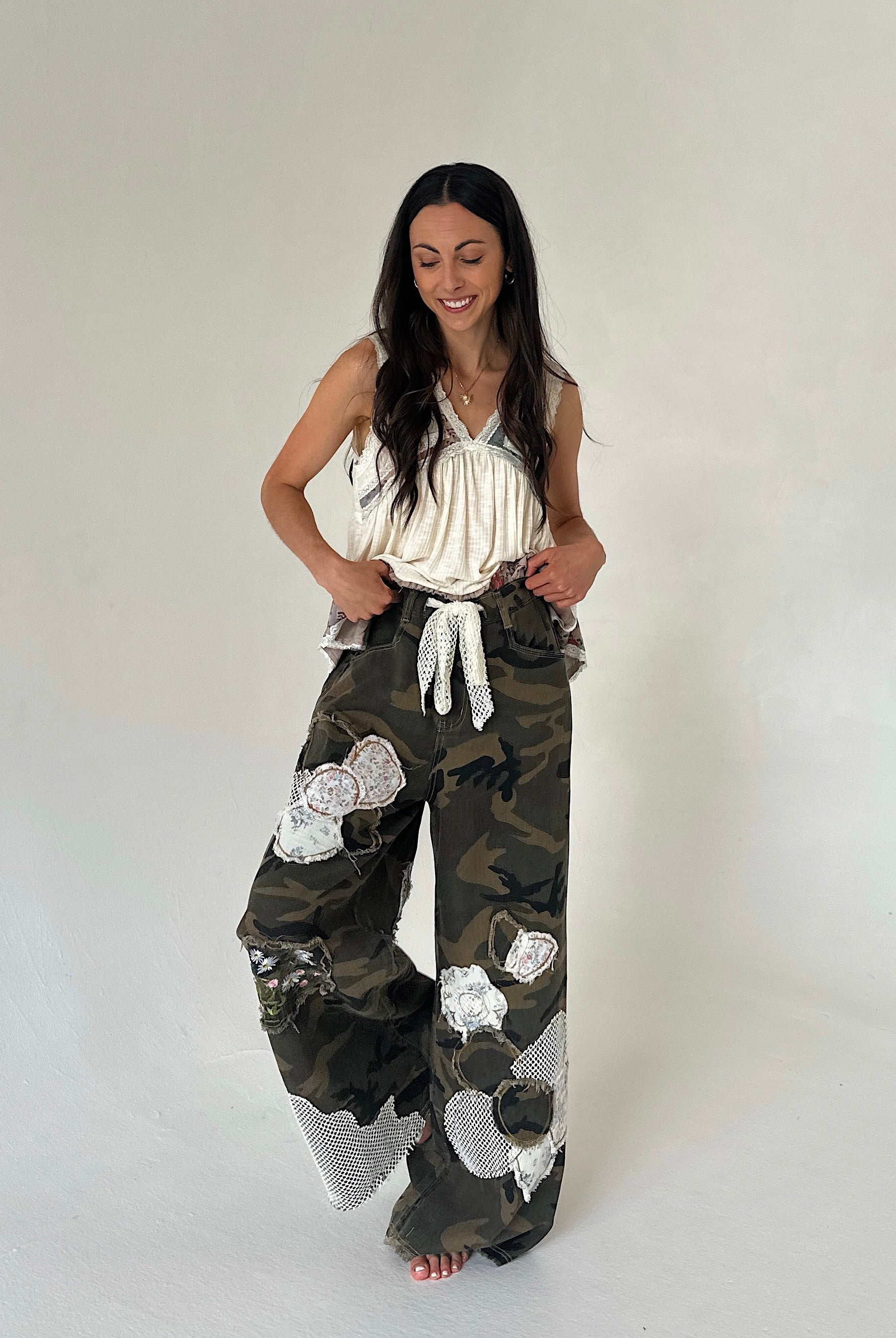 A woman modeling a pair of wide-leg camouflage pants with large white floral applique patches, paired with a cream sleeveless top, against a plain light gray studio background.