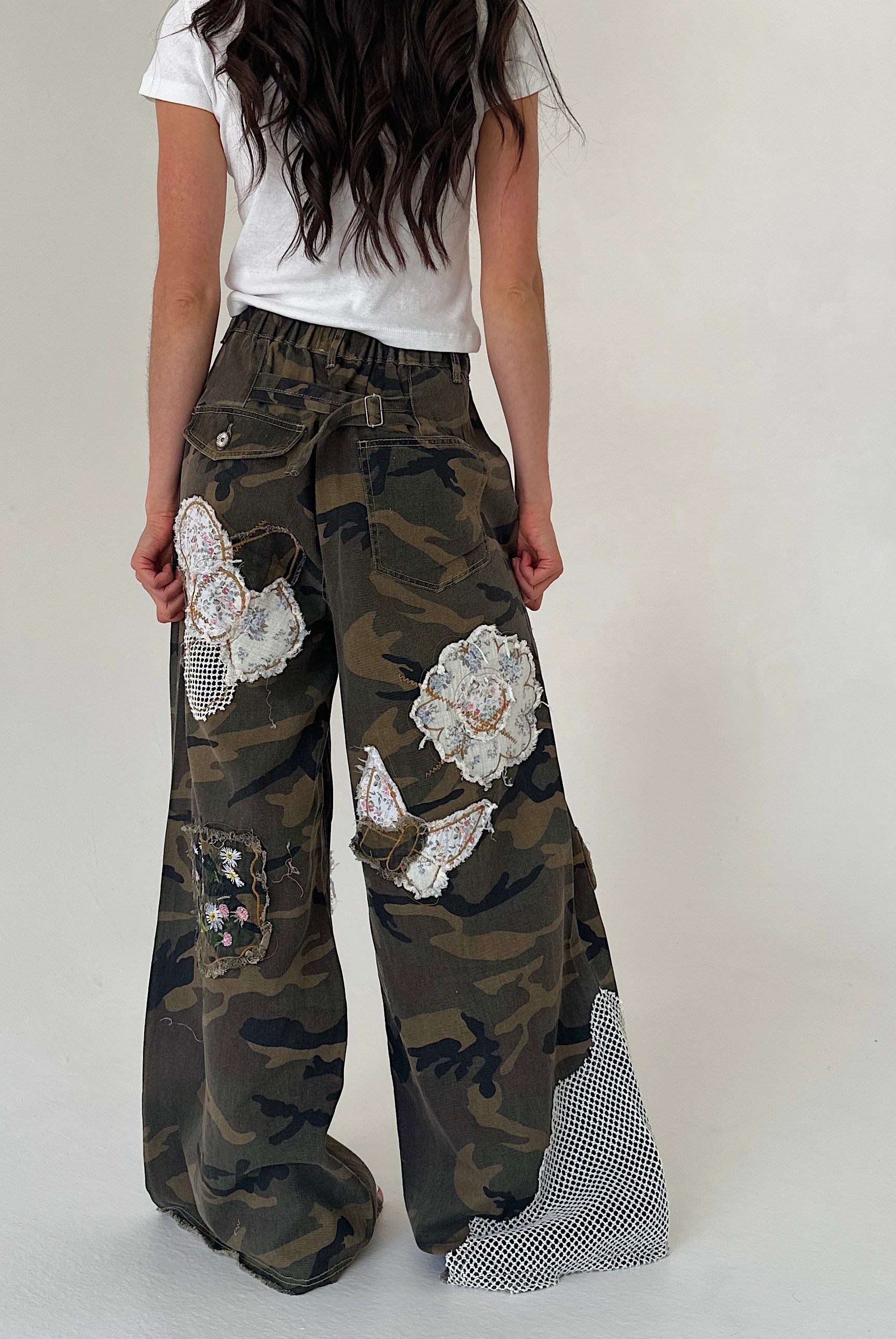 A person shown from the back wearing wide-leg camouflage pants with large white floral applique patches and a mesh fabric panel near the hem, paired with a white T-shirt against a plain light gray studio background.