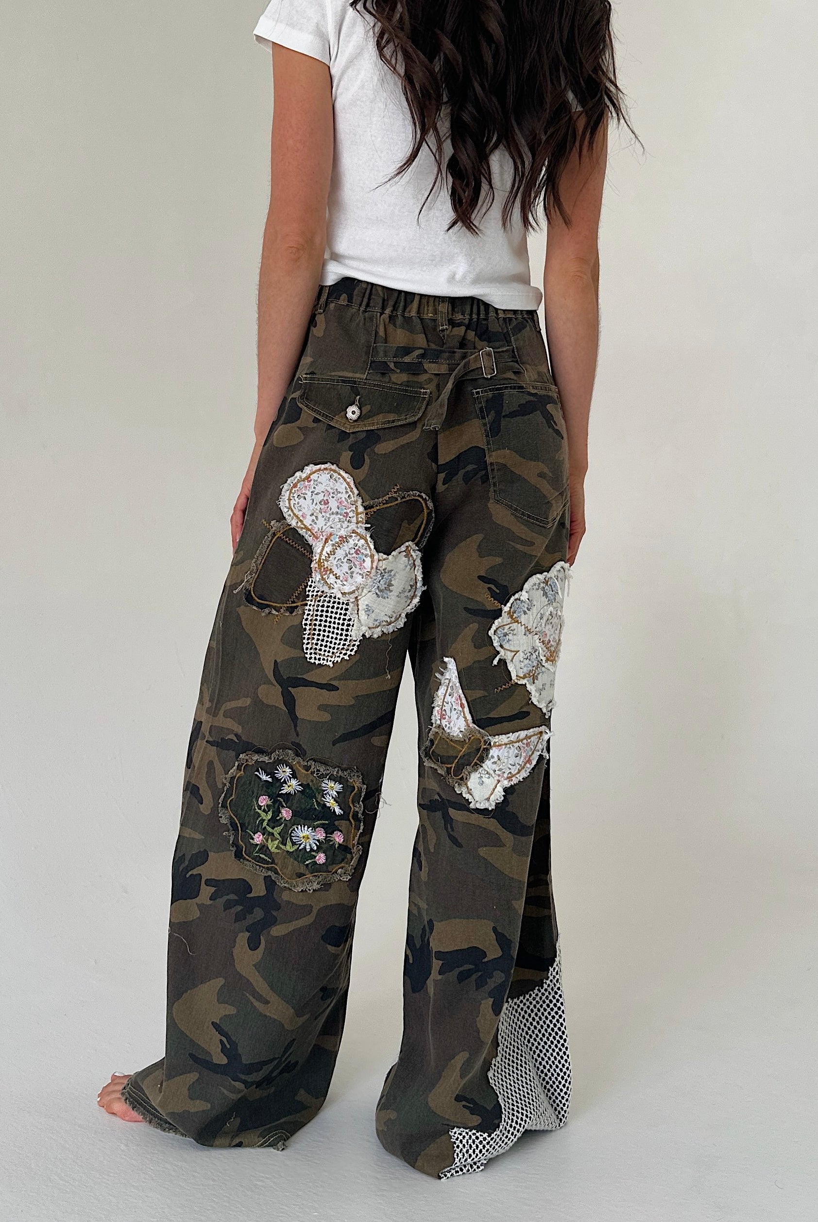 A person shown from the back wearing loose camouflage wide-leg pants with decorative white lace applique patches and a white top, against a plain light gray studio background.