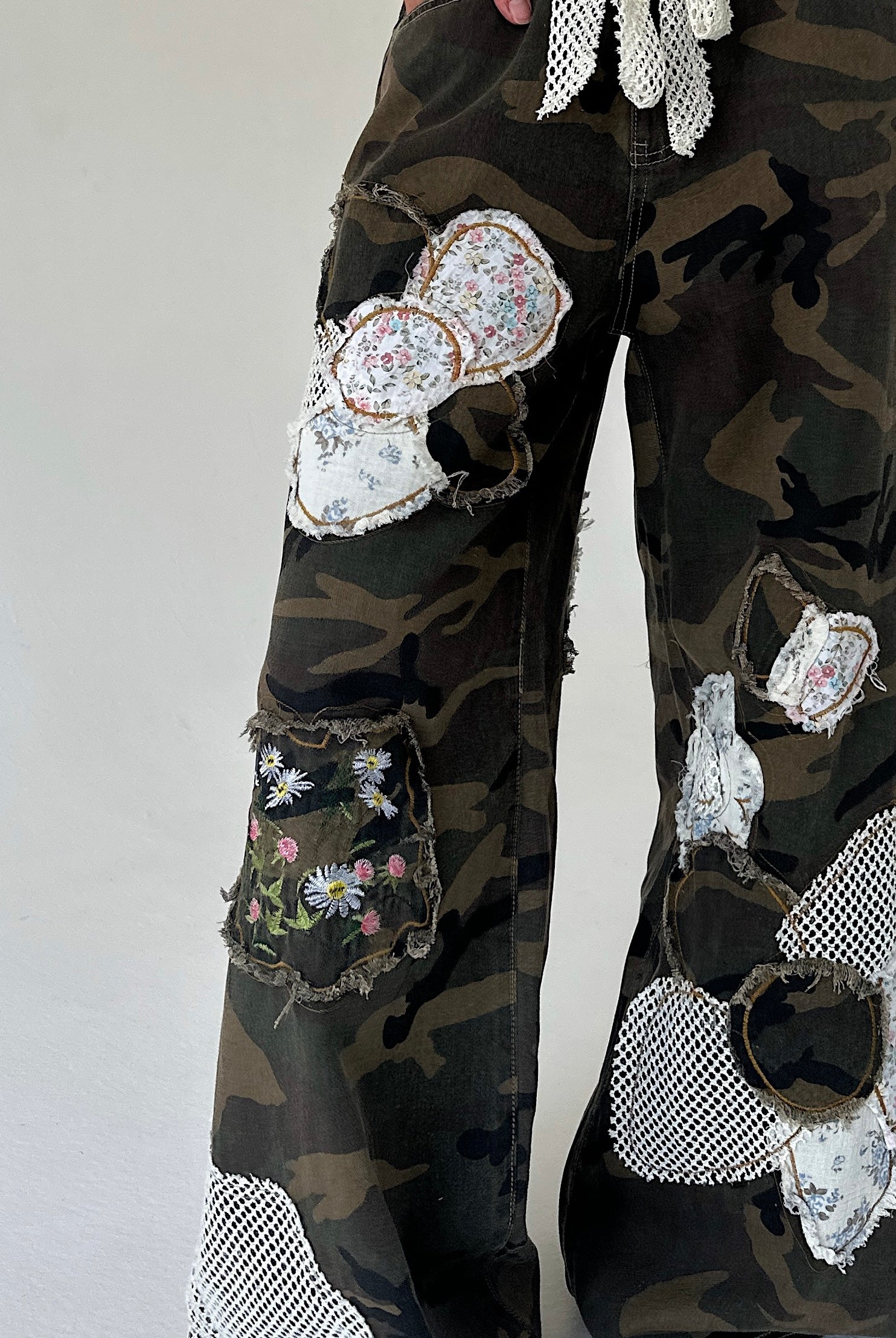 A close-up of a person's lower legs wearing camouflage wide-leg pants heavily embellished with patchwork floral fabric patches, mesh cutouts, and frayed details, set against a plain light gray background.