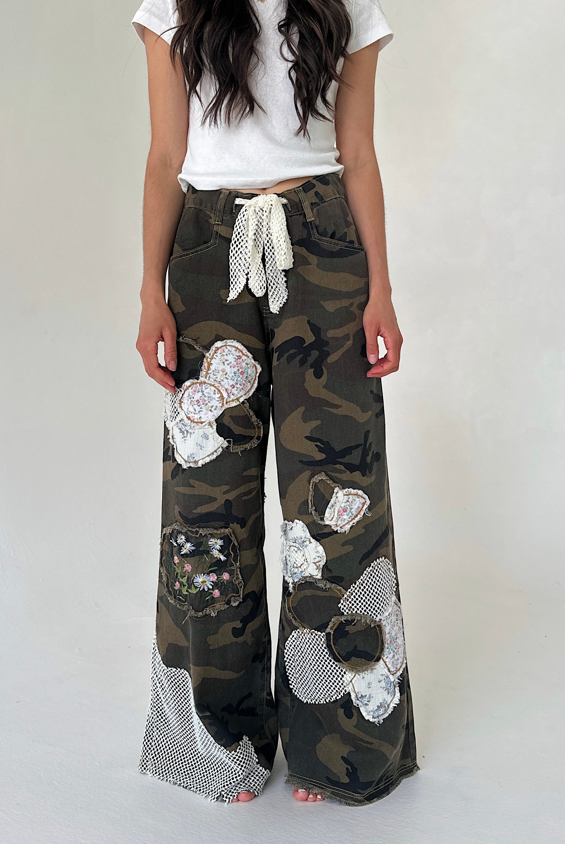 A person wearing loose camouflage wide-leg pants with large white flower appliqués and a white tie waist, paired with a white top, against a plain light background.