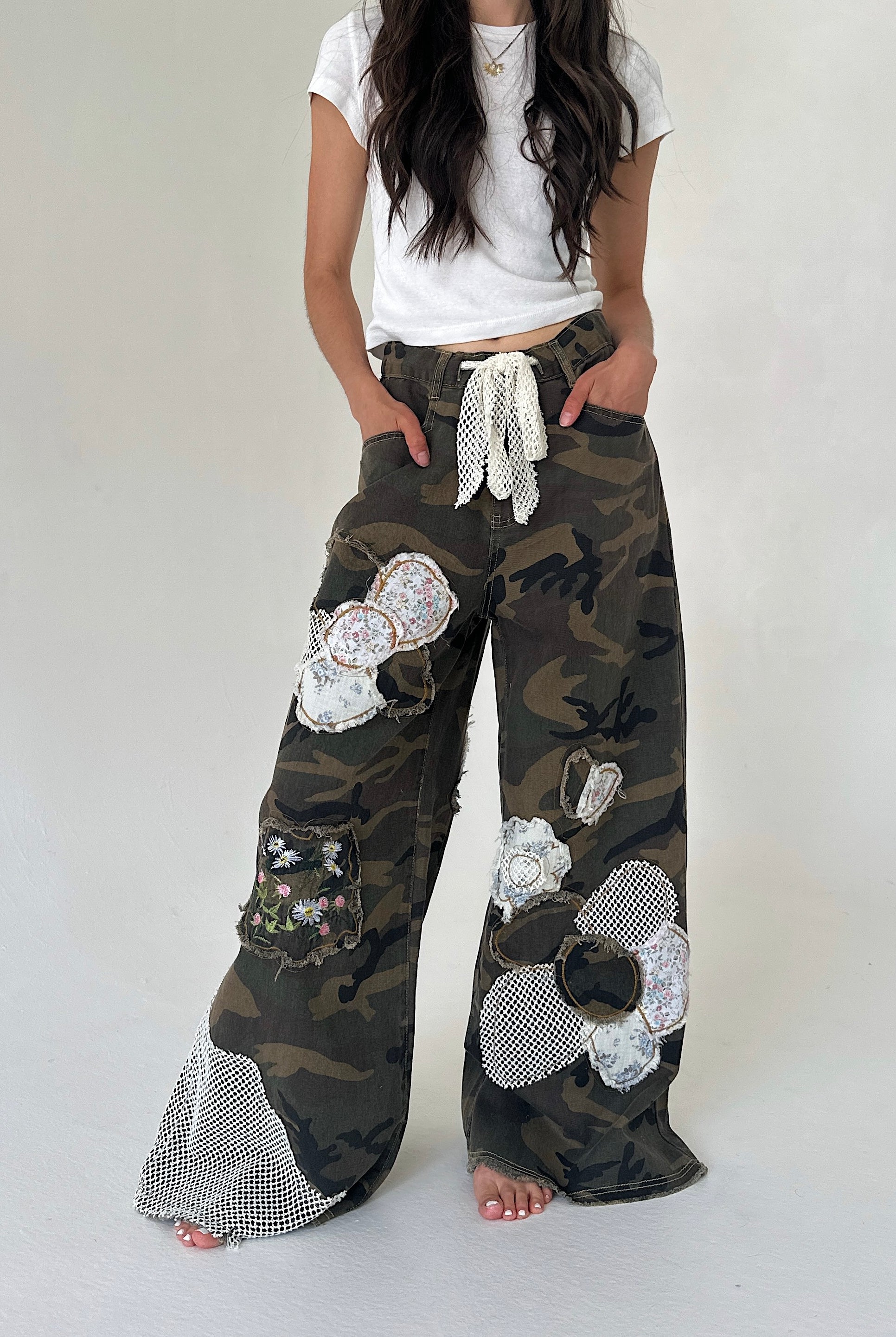 A person wearing loose camouflage wide-leg pants with large white sequined flower appliqués, a white drawstring waist, and mesh hem detailing, paired with a white cropped T-shirt against a plain light gray studio background.
