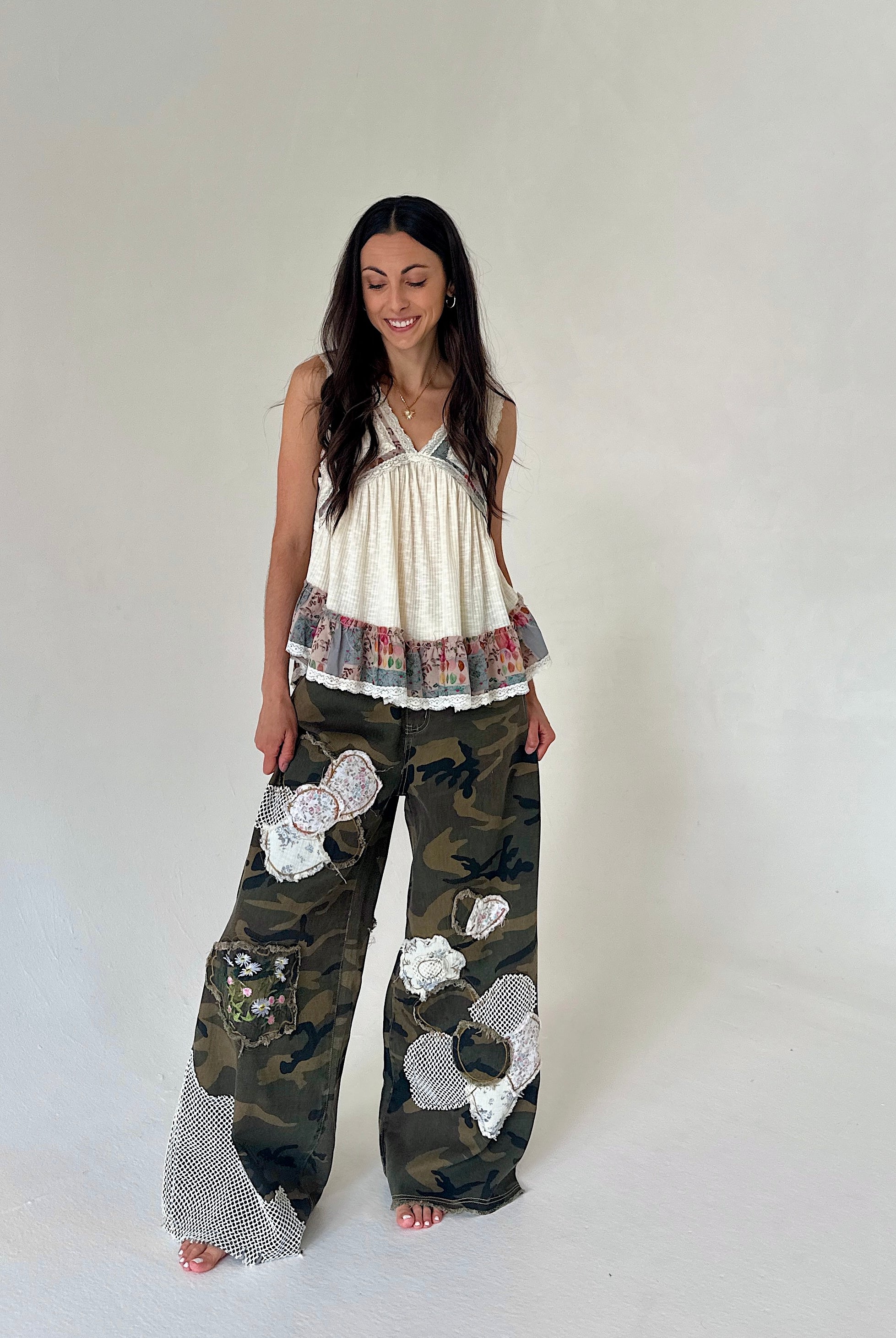 A woman modeling a sleeveless cream top with a ruffled hem and wide-leg camouflage pants featuring white mesh patchwork details, standing against a plain light gray studio background.