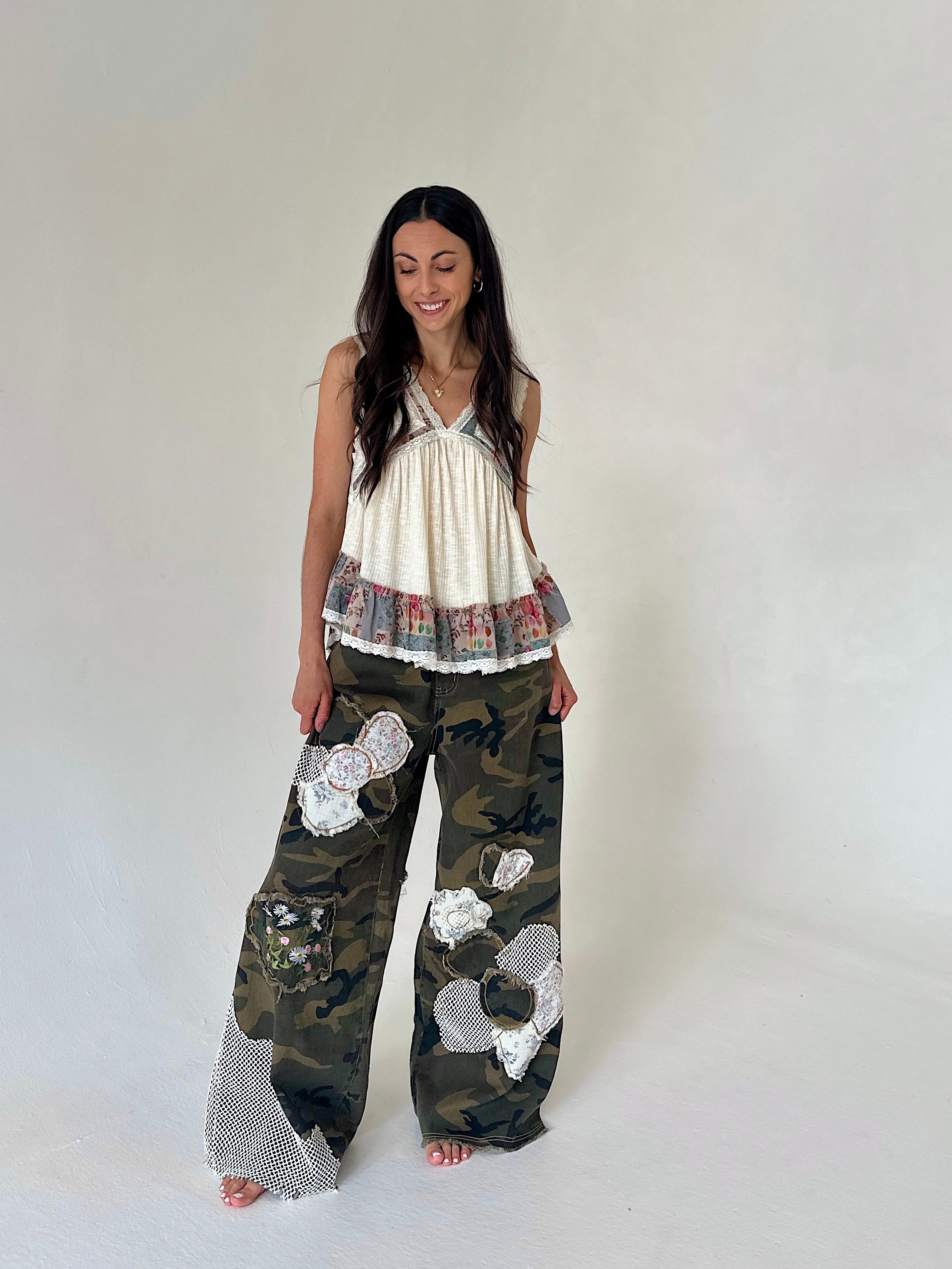 A woman modeling a sleeveless cream top with a ruffled hem and wide-leg camouflage pants featuring white mesh patchwork details, standing against a plain light gray studio background.