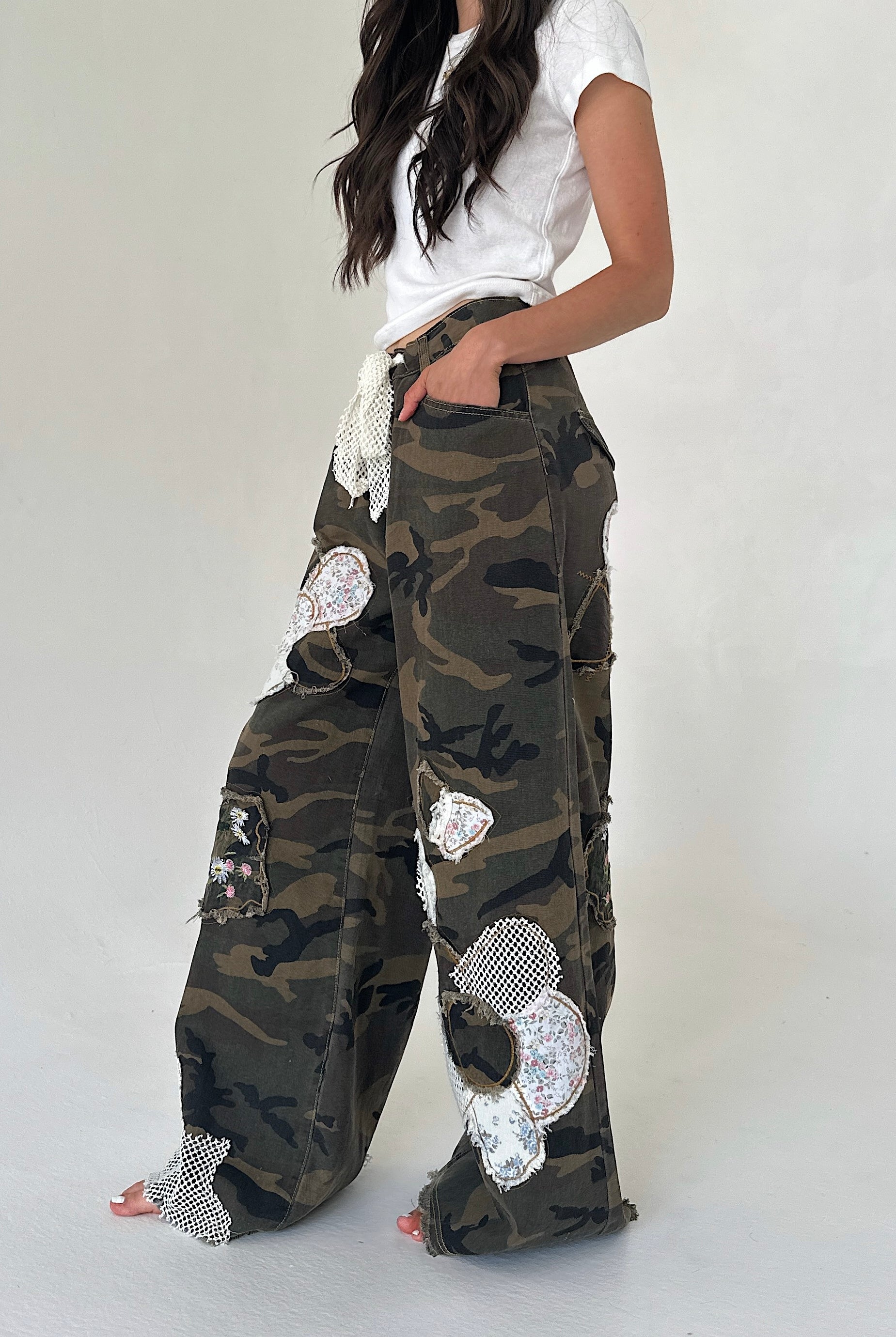 A model wearing a pair of wide-leg camouflage cargo pants decorated with floral patches and mesh trim, paired with a white t-shirt against a plain light gray background.
