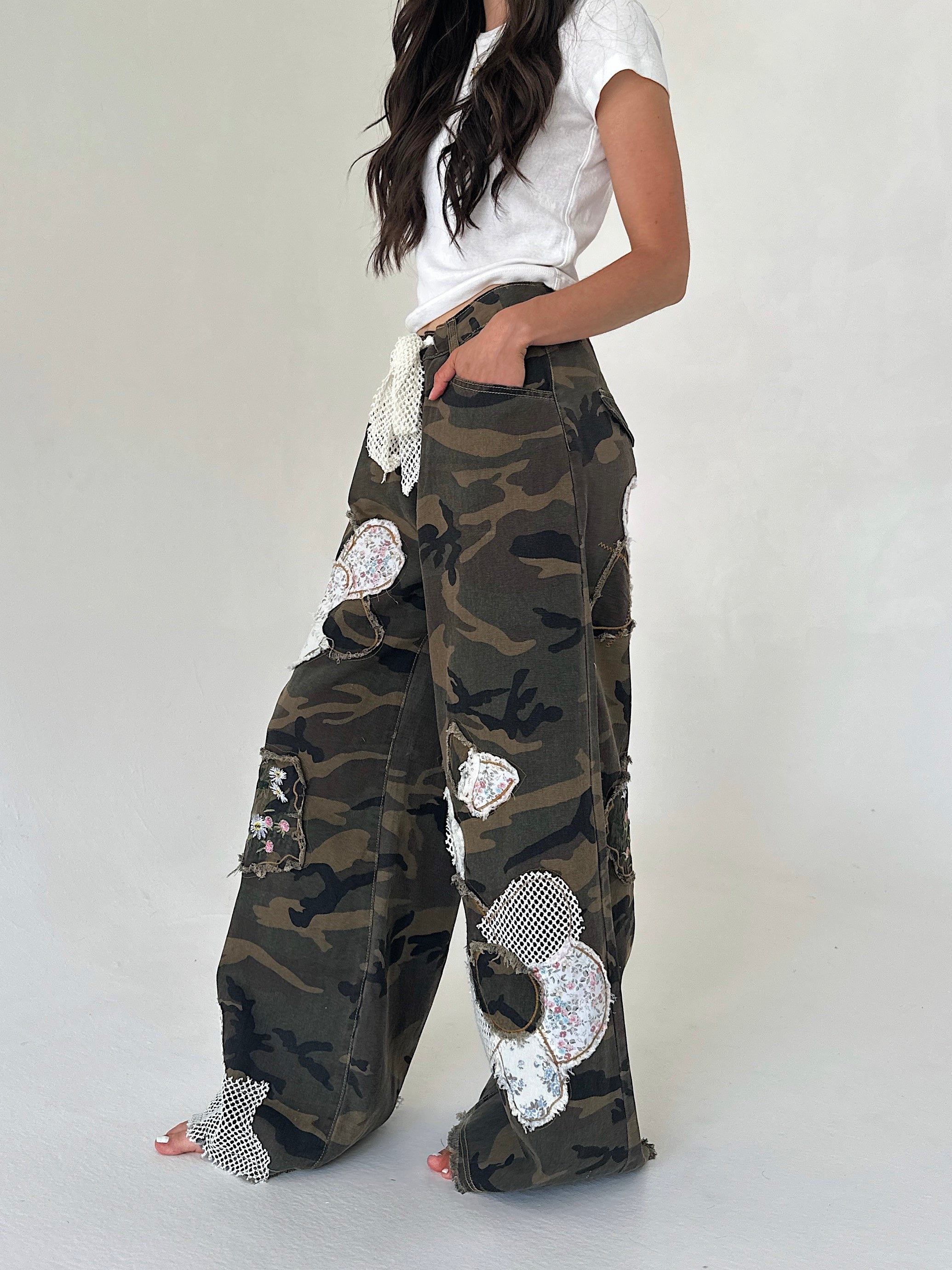 A model wearing a pair of wide-leg camouflage cargo pants decorated with floral patches and mesh trim, paired with a white t-shirt against a plain light gray background.