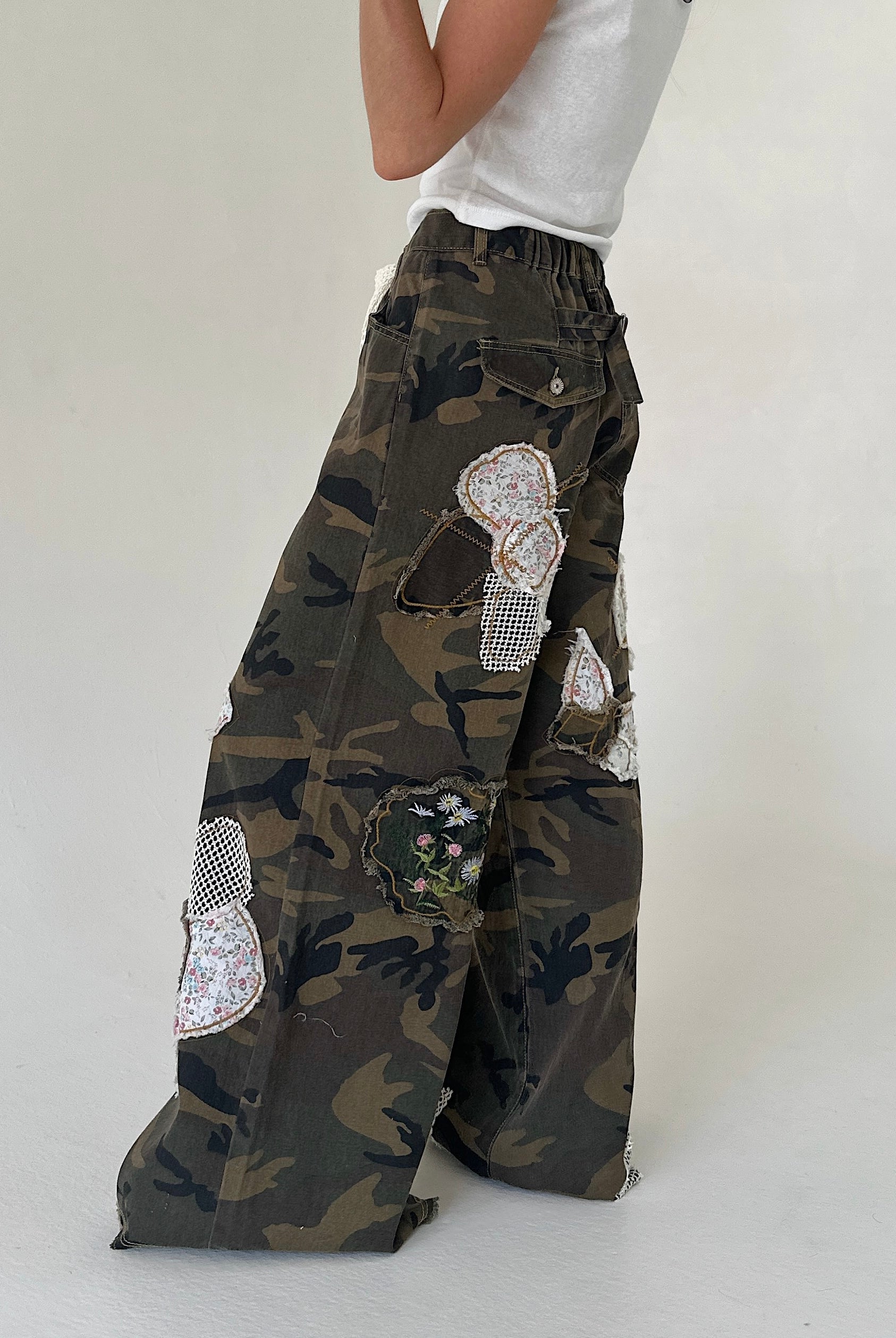 A person wearing wide-leg camouflage cargo pants with large decorative floral patches, paired with a white sleeveless top, photographed against a plain light gray studio background.