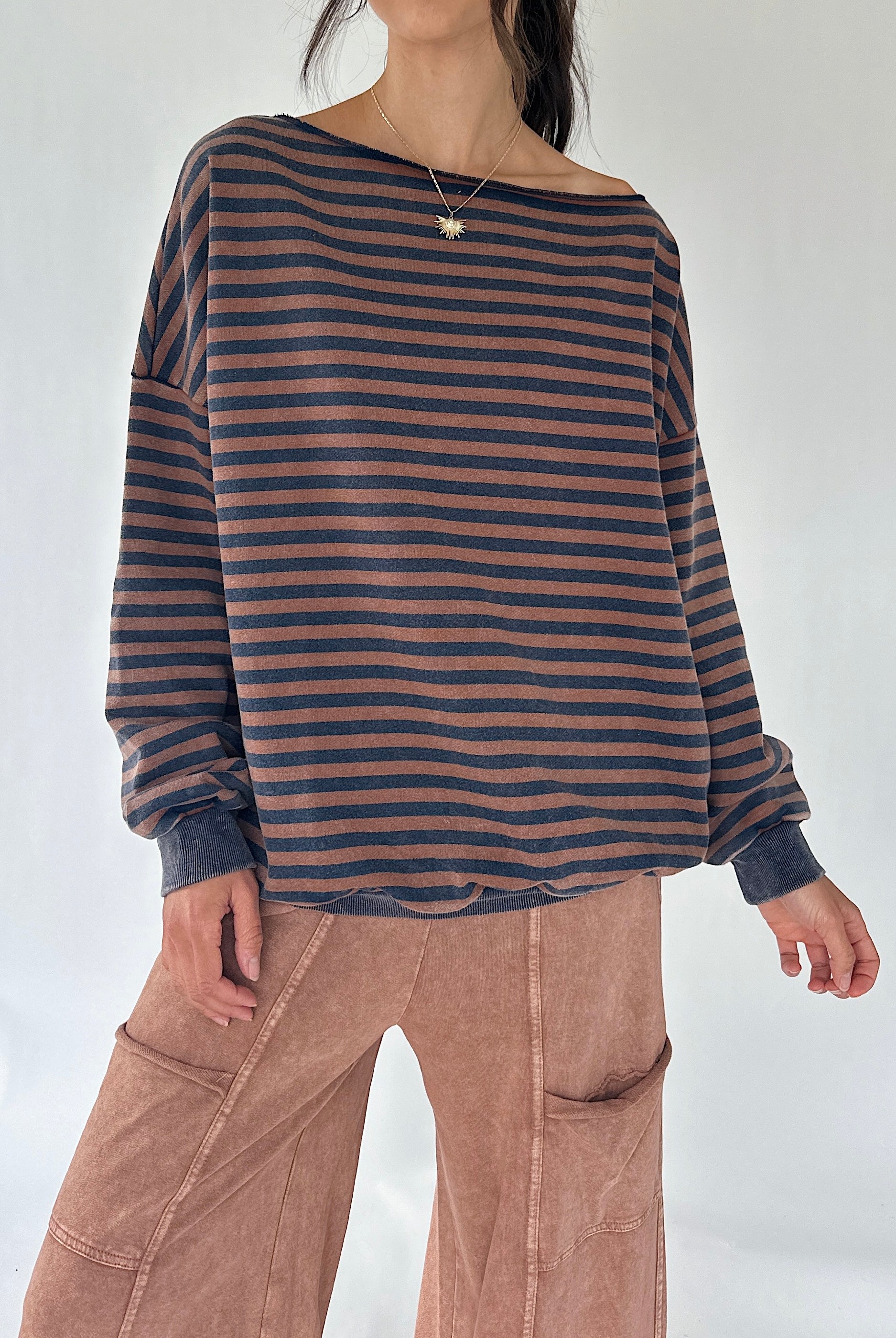 A woman wearing a loose, off-the-shoulder long-sleeve top with horizontal dark brown and navy stripes, paired with dusty rose wide-leg pants, against a plain light background.