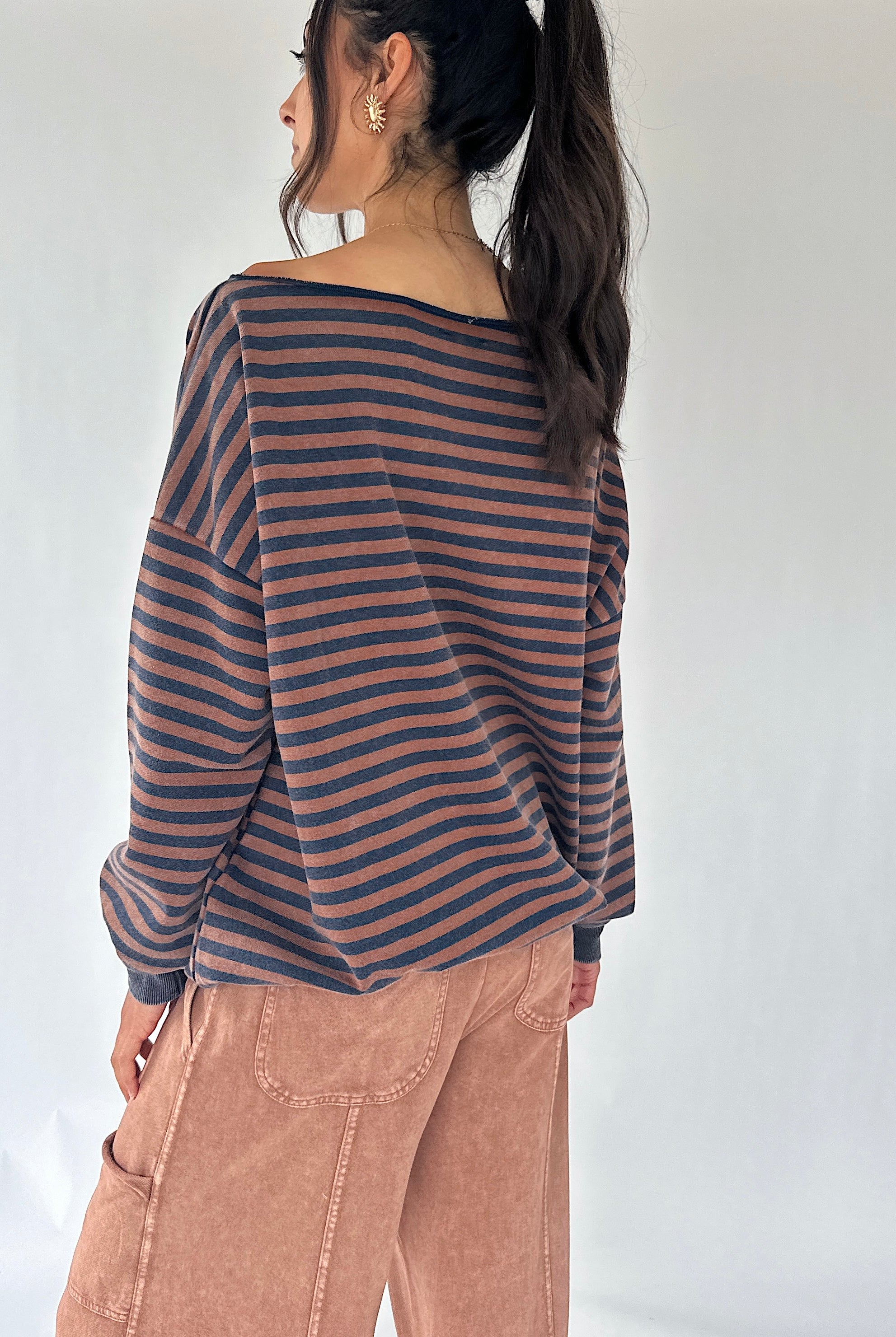 A woman shown from the back wearing a loose, off-the-shoulder long-sleeve top with horizontal dark brown and dusty navy stripes, paired with dusty brown wide leg pants, against a plain white studio background.