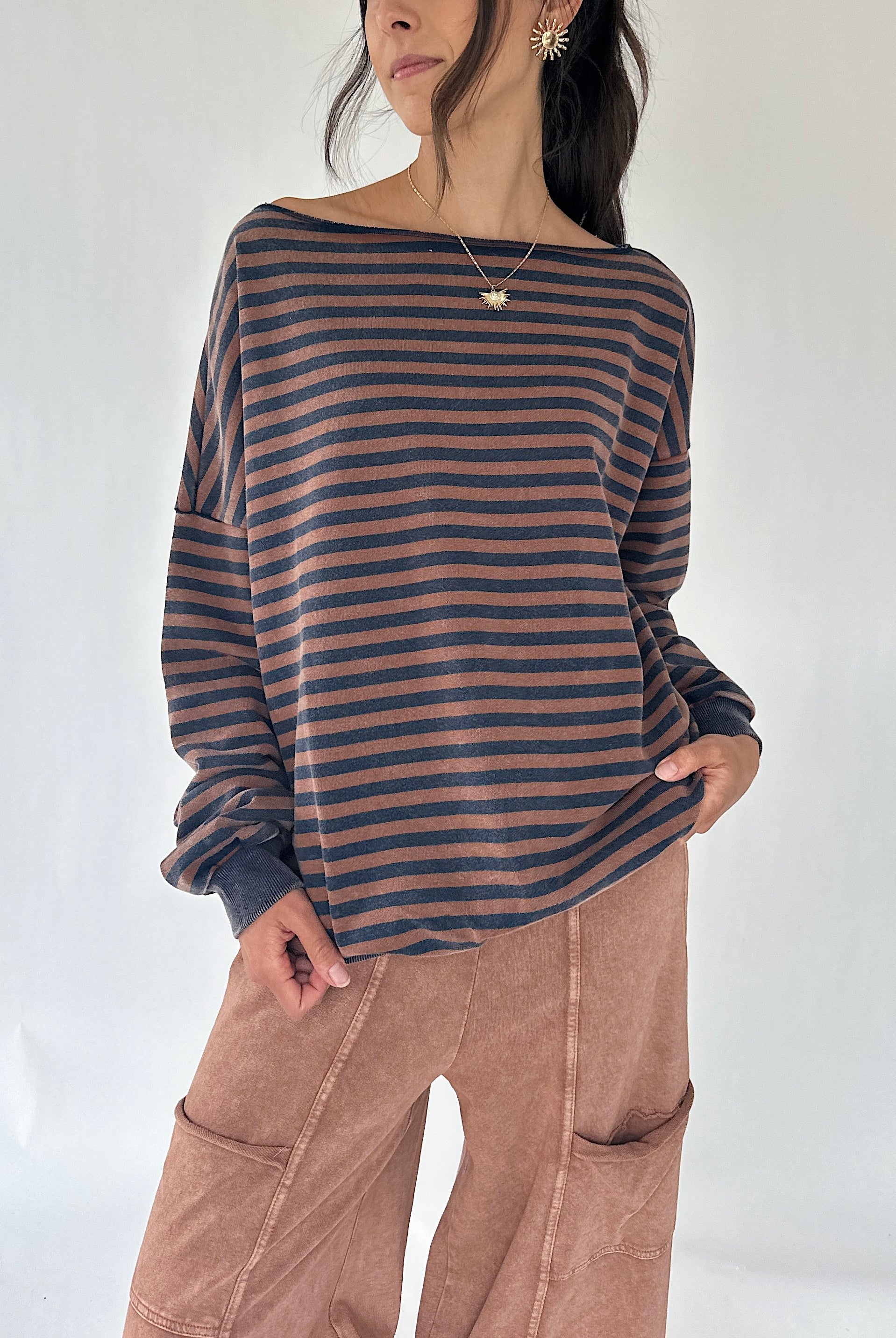 A woman wearing a loose, off-the-shoulder long-sleeve top with horizontal dark brown and muted blue stripes, paired with brown wide-leg pants, against a plain white studio background.