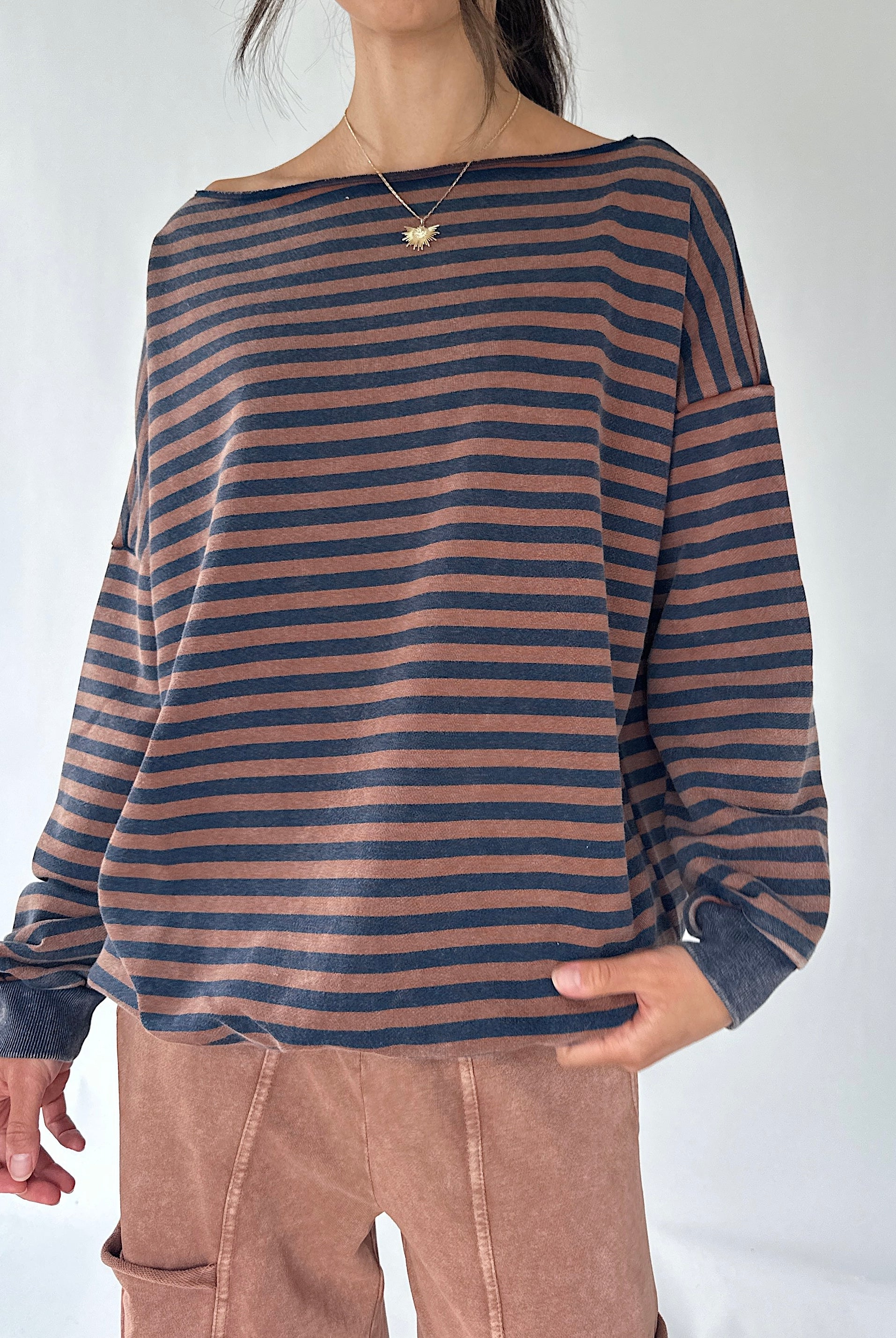 A woman wearing a loose, long-sleeve striped top with horizontal navy and dusty rose-brown stripes, paired with brown pants, against a plain light background.