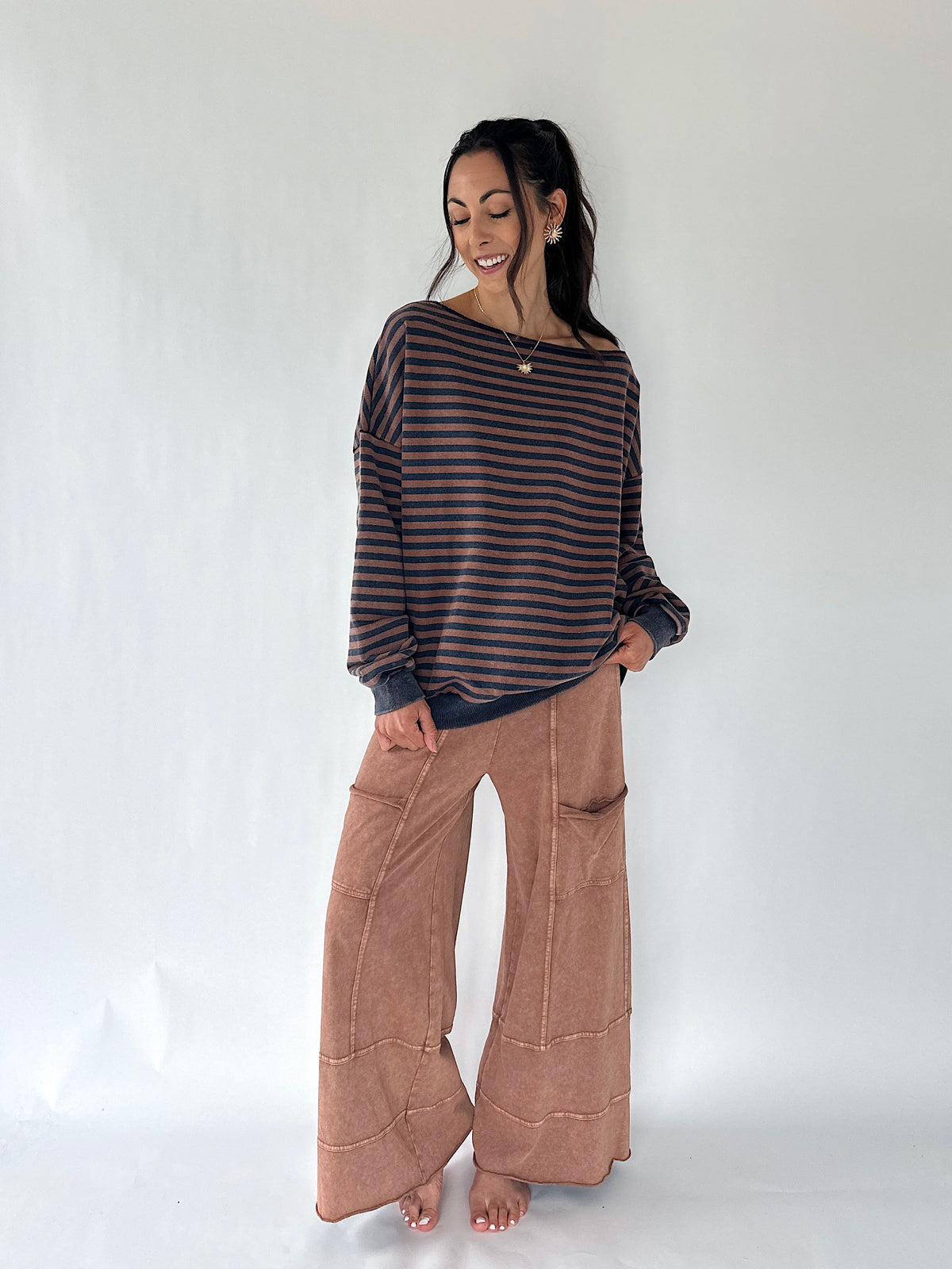 A woman modeling a dark brown and black striped long-sleeve knit top with wide-leg brown pants, standing against a plain white studio background.