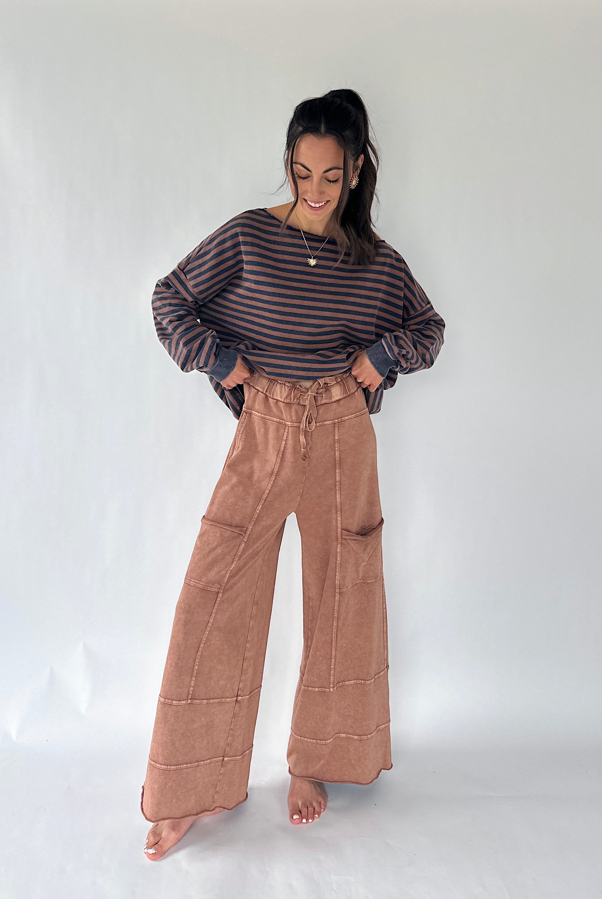 A woman stands barefoot against a plain light gray studio background, wearing a dark striped long-sleeve top and high-waisted wide-leg tan pants.
