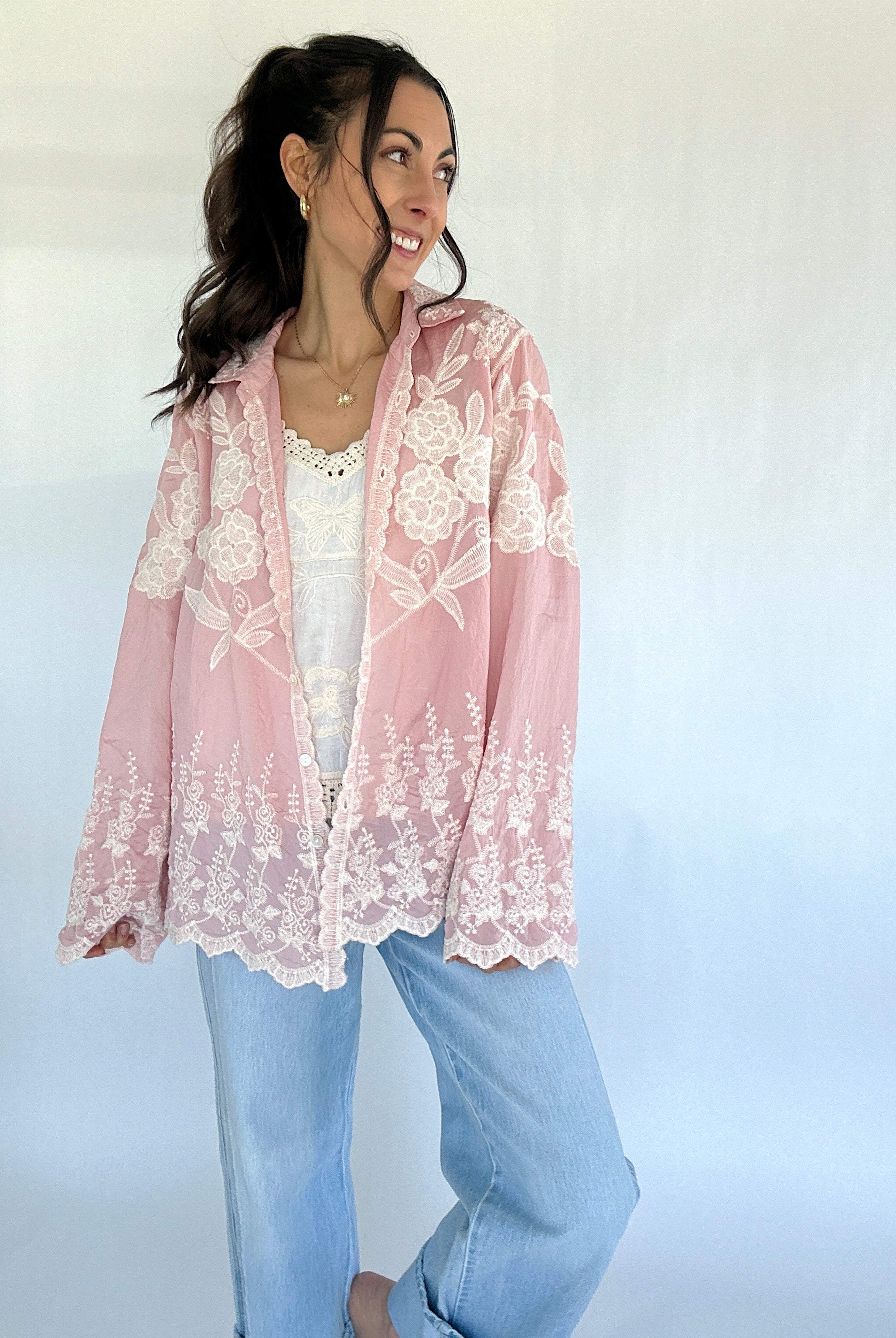 Woman wearing a pink lace button down over a white top and blue jeans on a light background