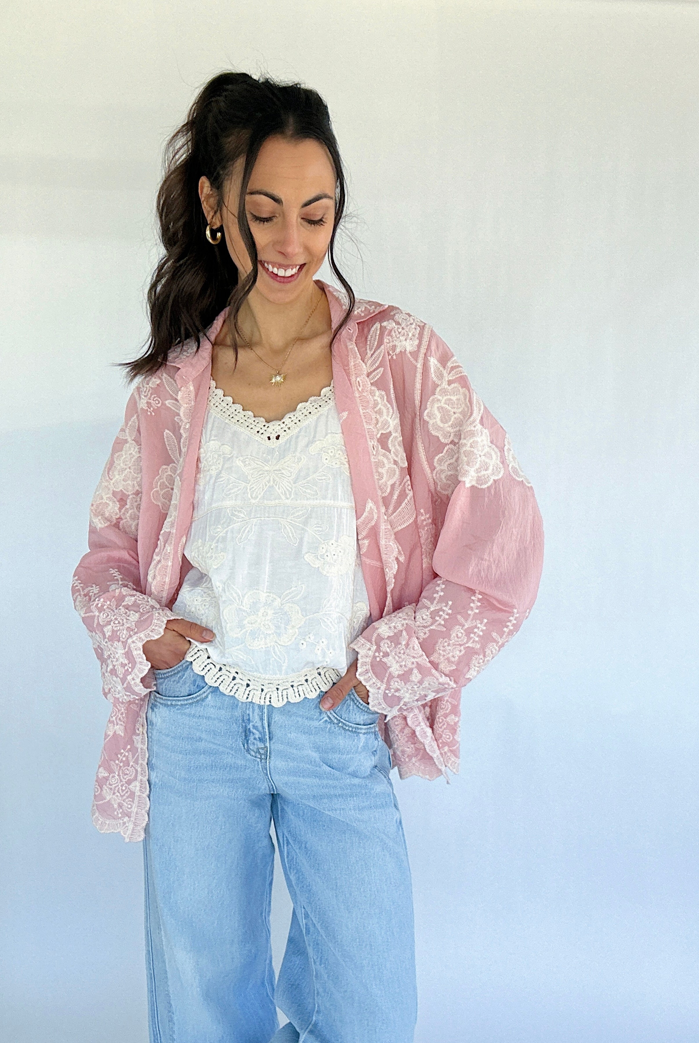 Woman wearing a pink lace long sleeve over a white top and blue jeans against a white background