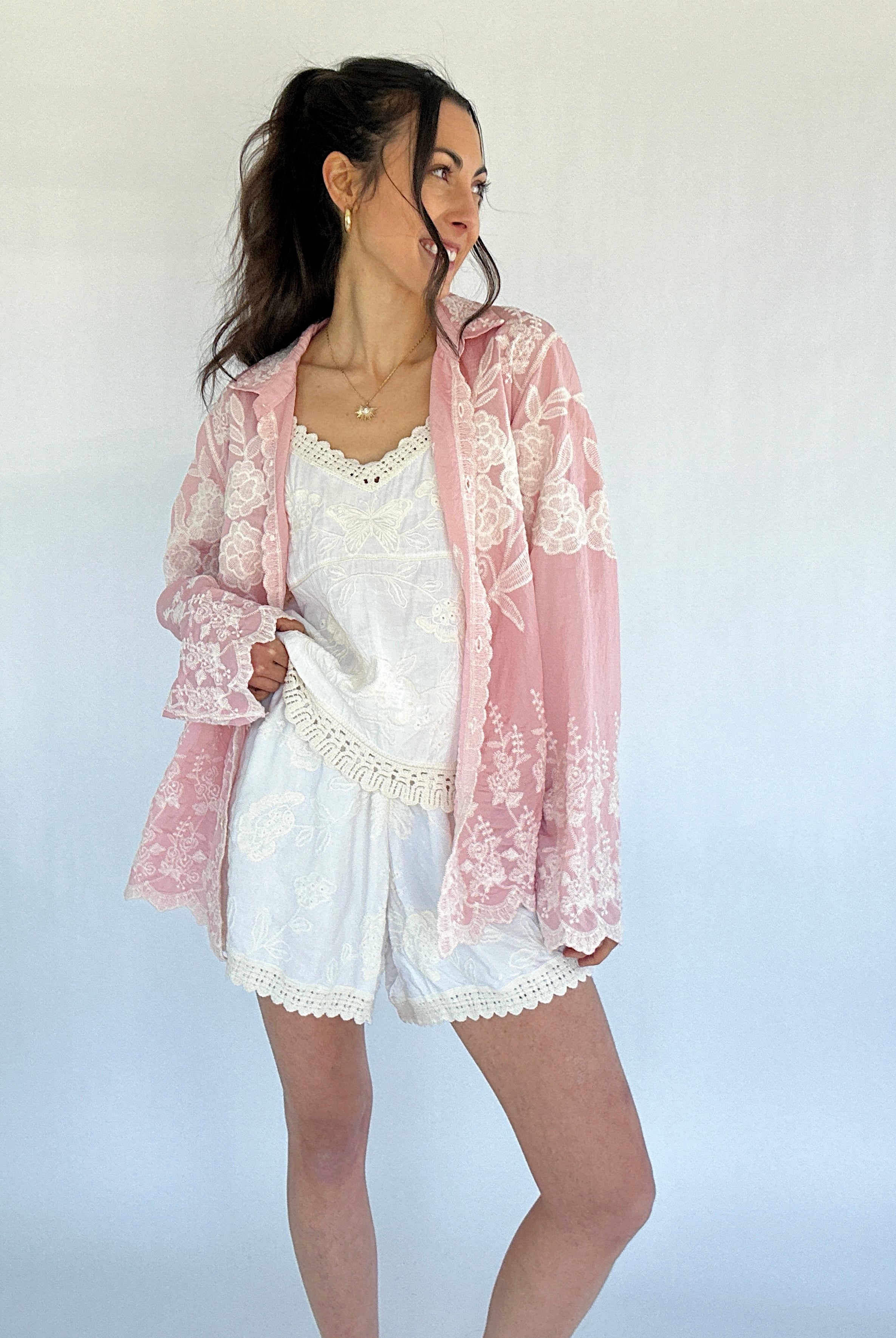 Woman wearing a pink lace long sleeve top over a white outfit on a plain background