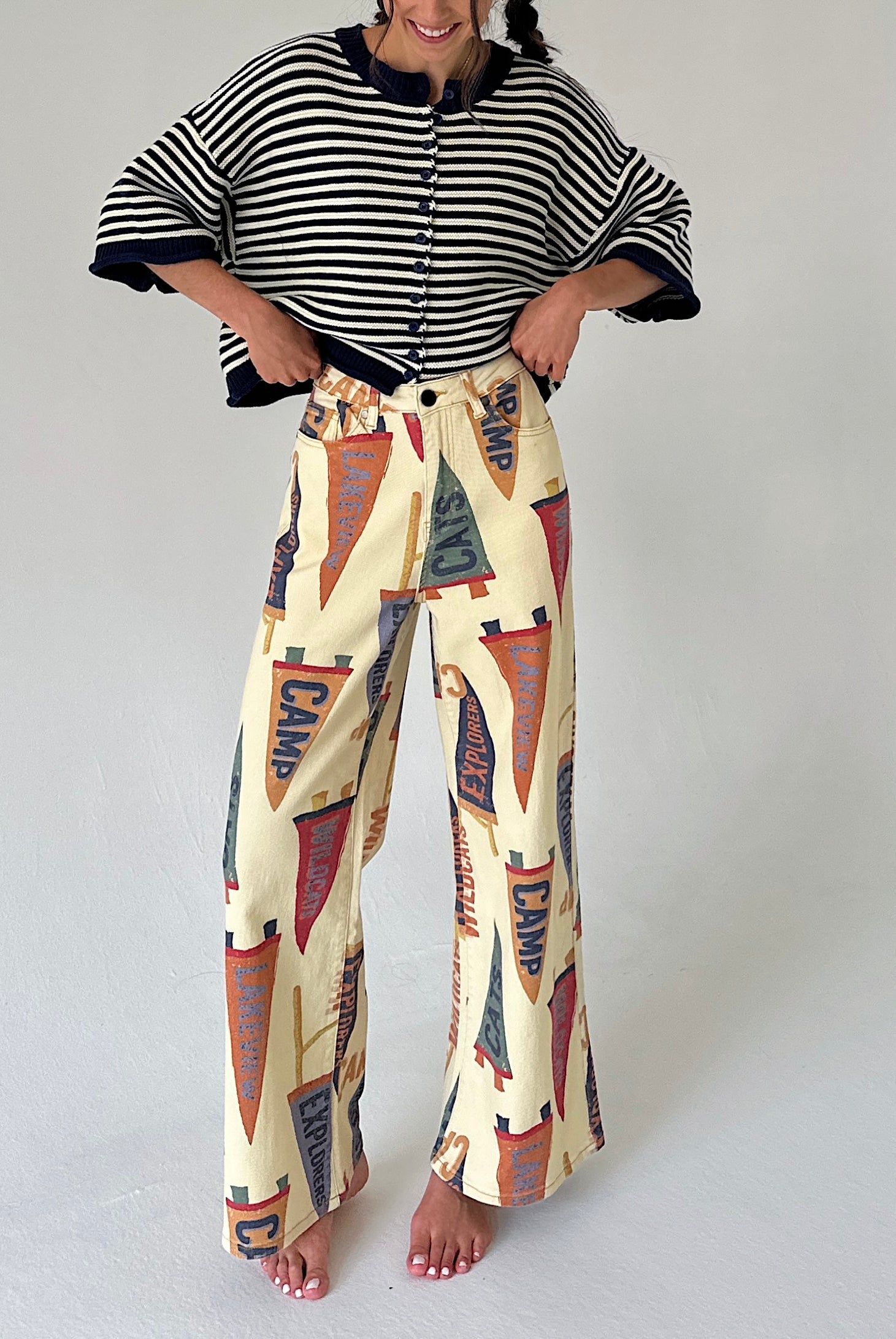 A woman modeling a two-piece outfit consisting of a blue and white striped short-sleeve cardigan and high-waisted, wide-leg pants featuring a vintage pennant flag print on a cream background.