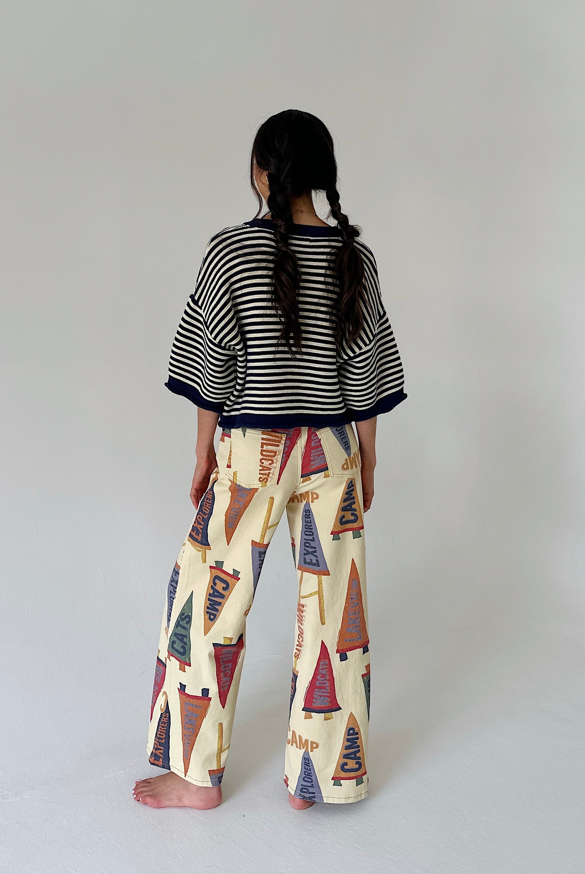 A child model shown from behind wearing a black and white striped short-sleeve top and cream wide-leg pants with a colorful pennant flag pattern, standing against a plain light gray studio background.